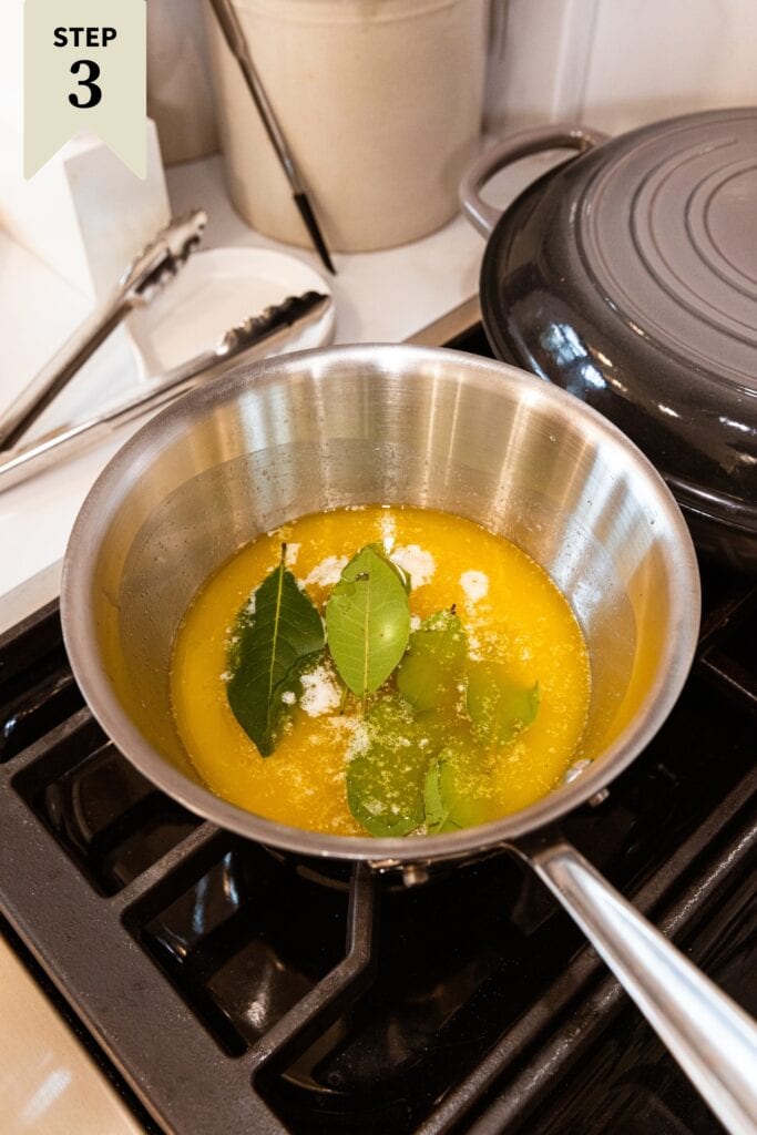 Saucepan sitting on stovetop with melting white wine, butter, and bay leaves mixture.