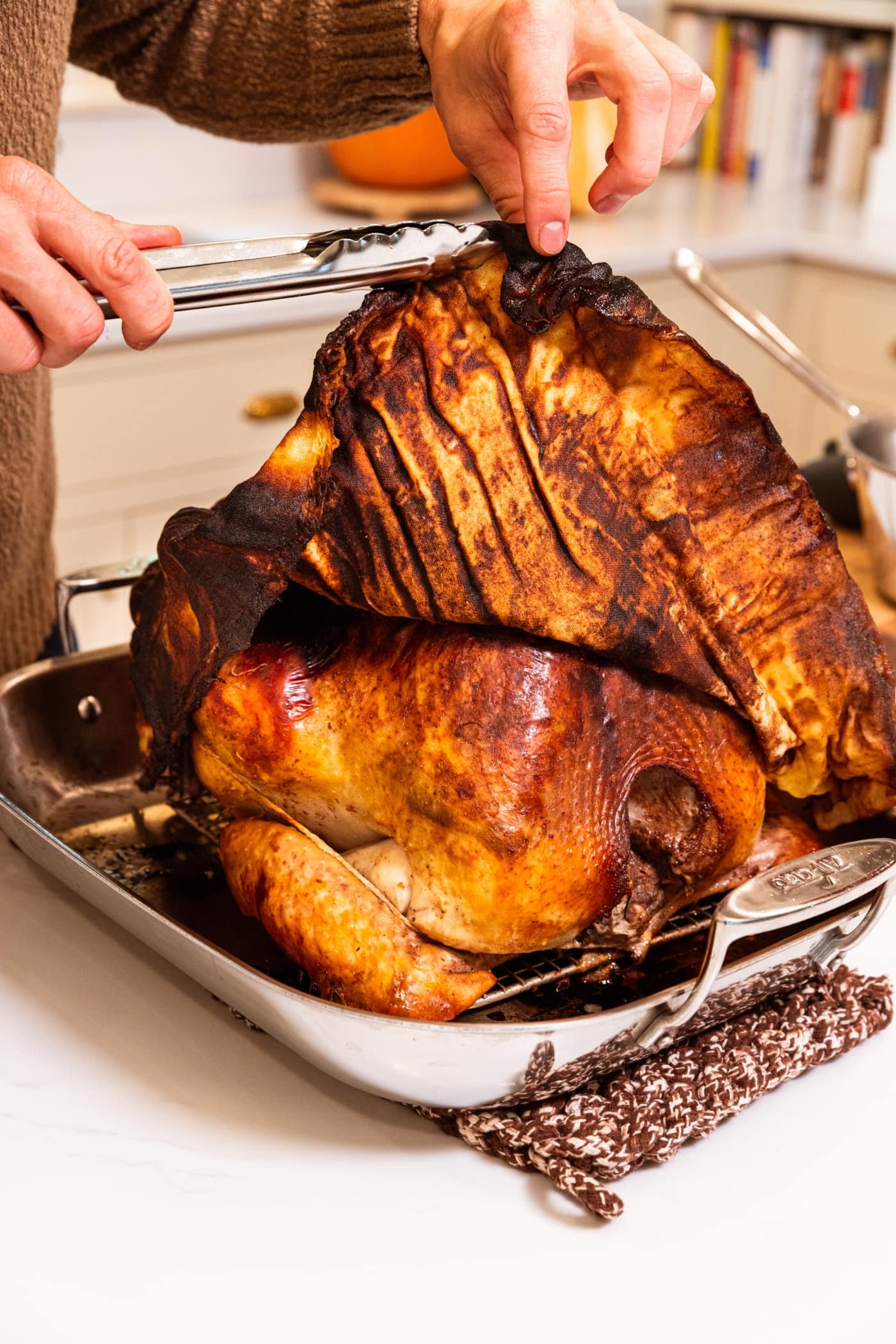 Two hands pulling back cheesecloth from a large Thanksgiving turkey showing golden texture underneath.