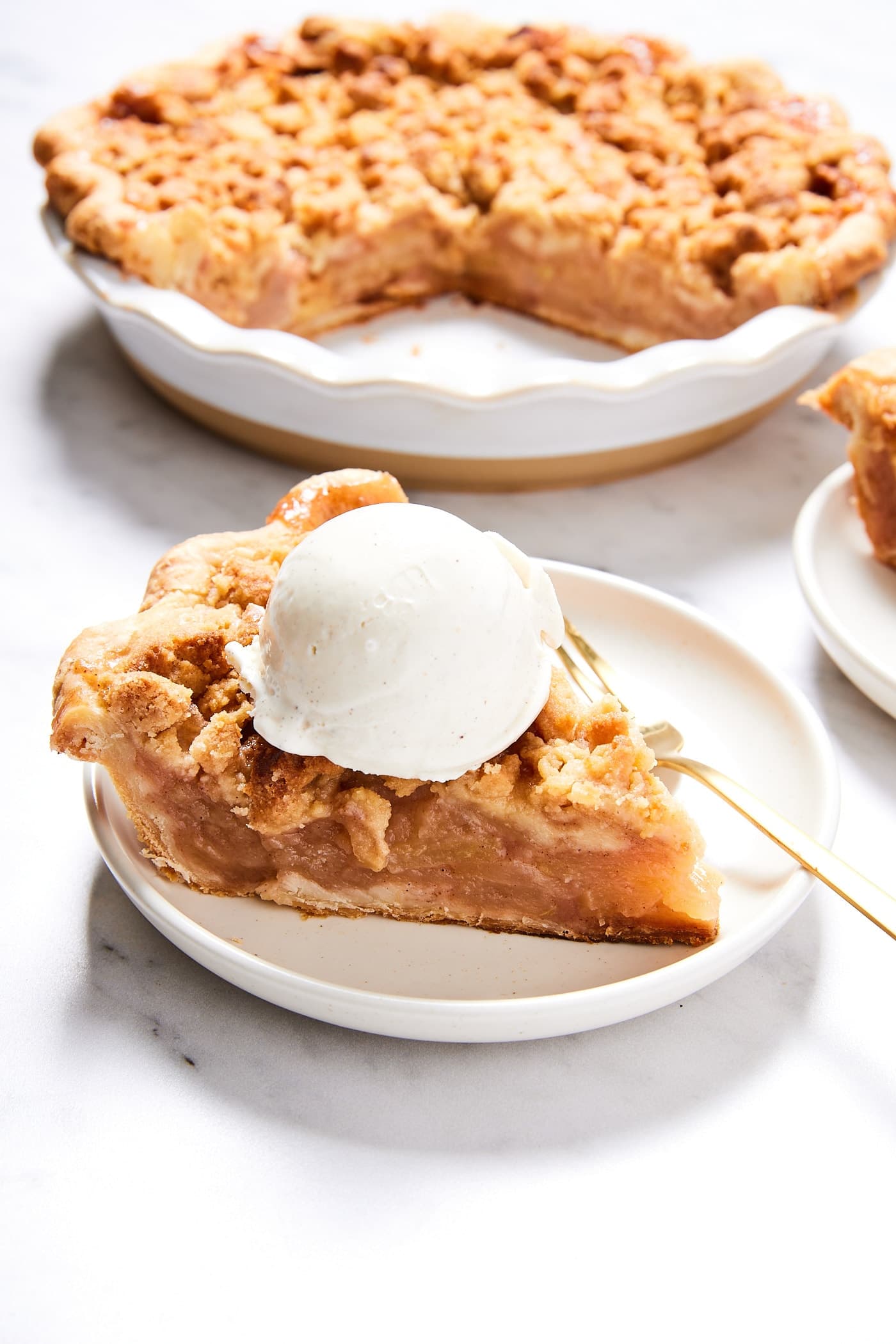White plate with slice of classic apple pie with scoop of ice cream on top with rest of the pie in the background.