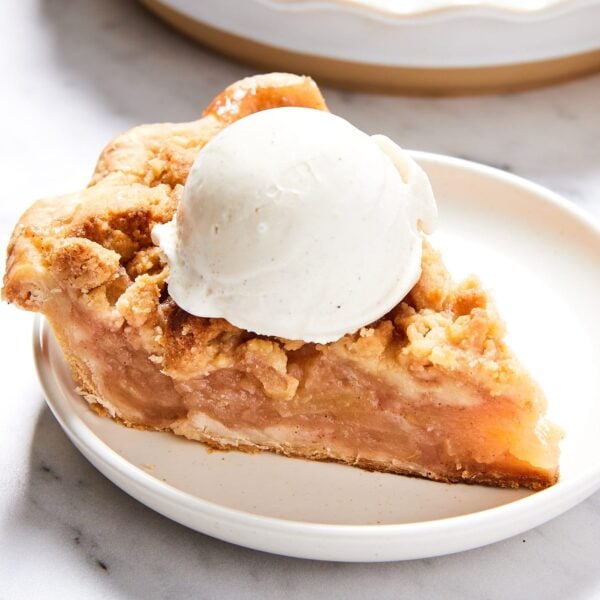 White plate with slice of classic apple pie with scoop of vanilla ice cream on top with rest of pie in the background.