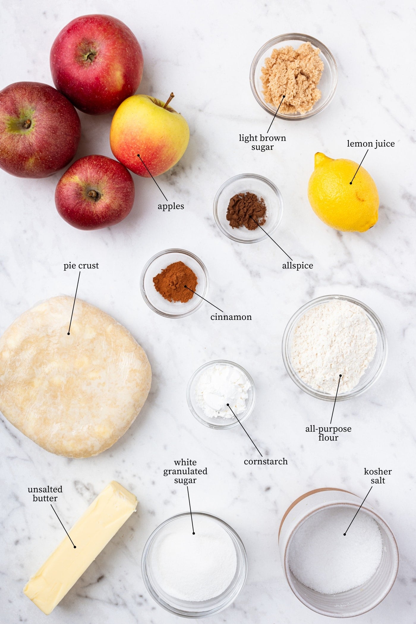 White marble surface filled with all ingredients needed to make an apple pie including apples, sugar, butter, spices, and more.