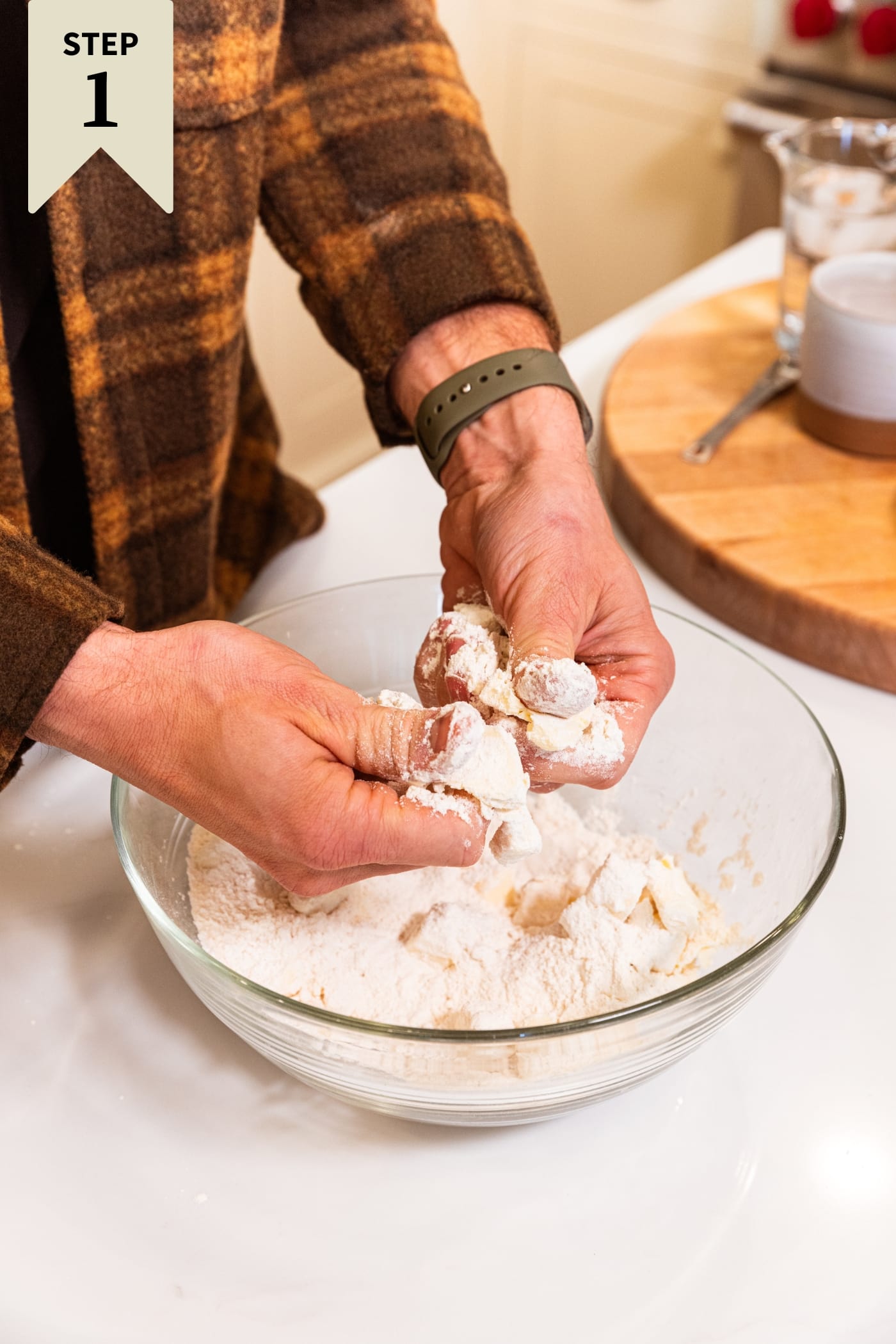 Two hands pressing together pie dough in glass bowl before it comes together fully.