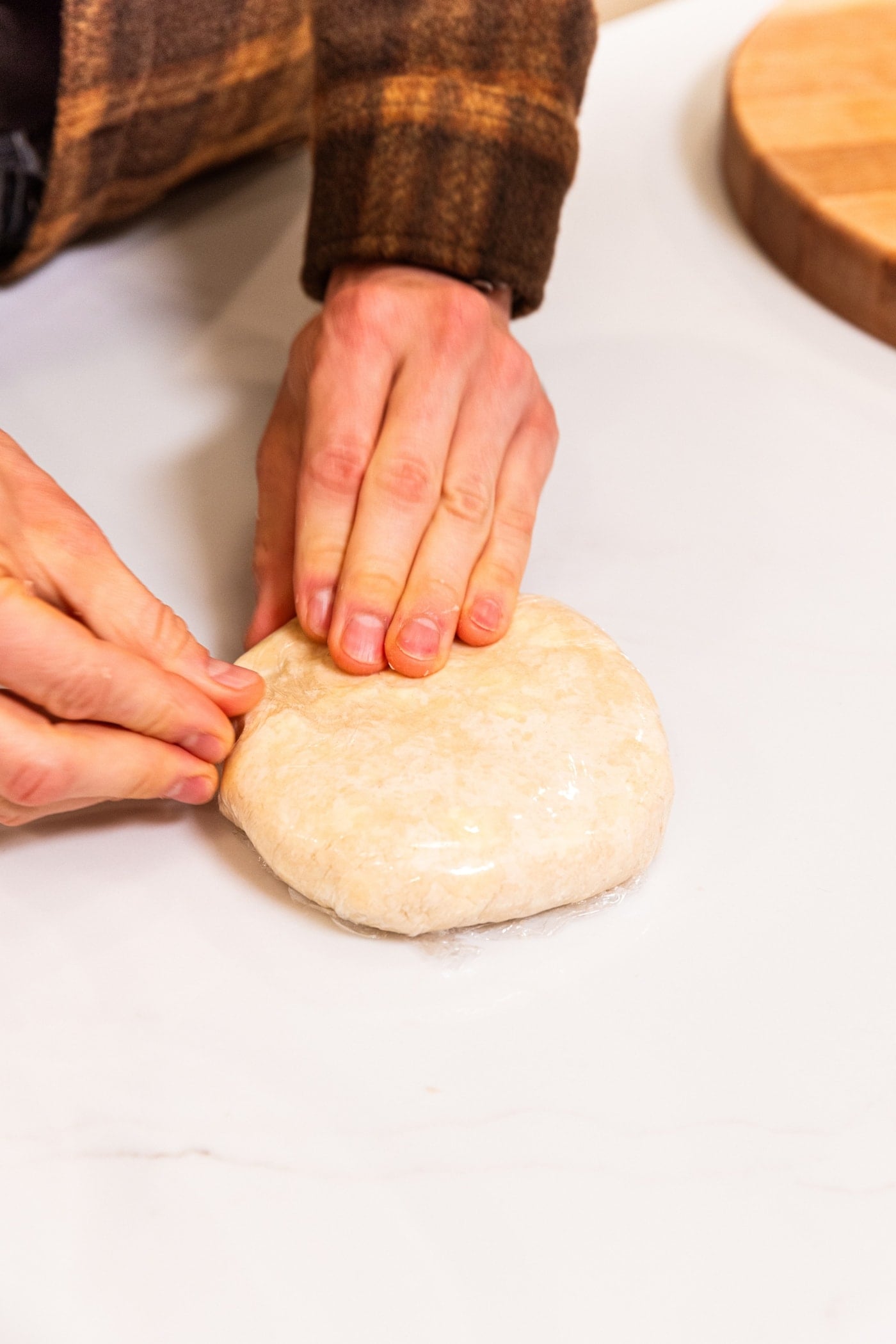 Hands pressing pie dough into a round in plastic wrap on white surface.