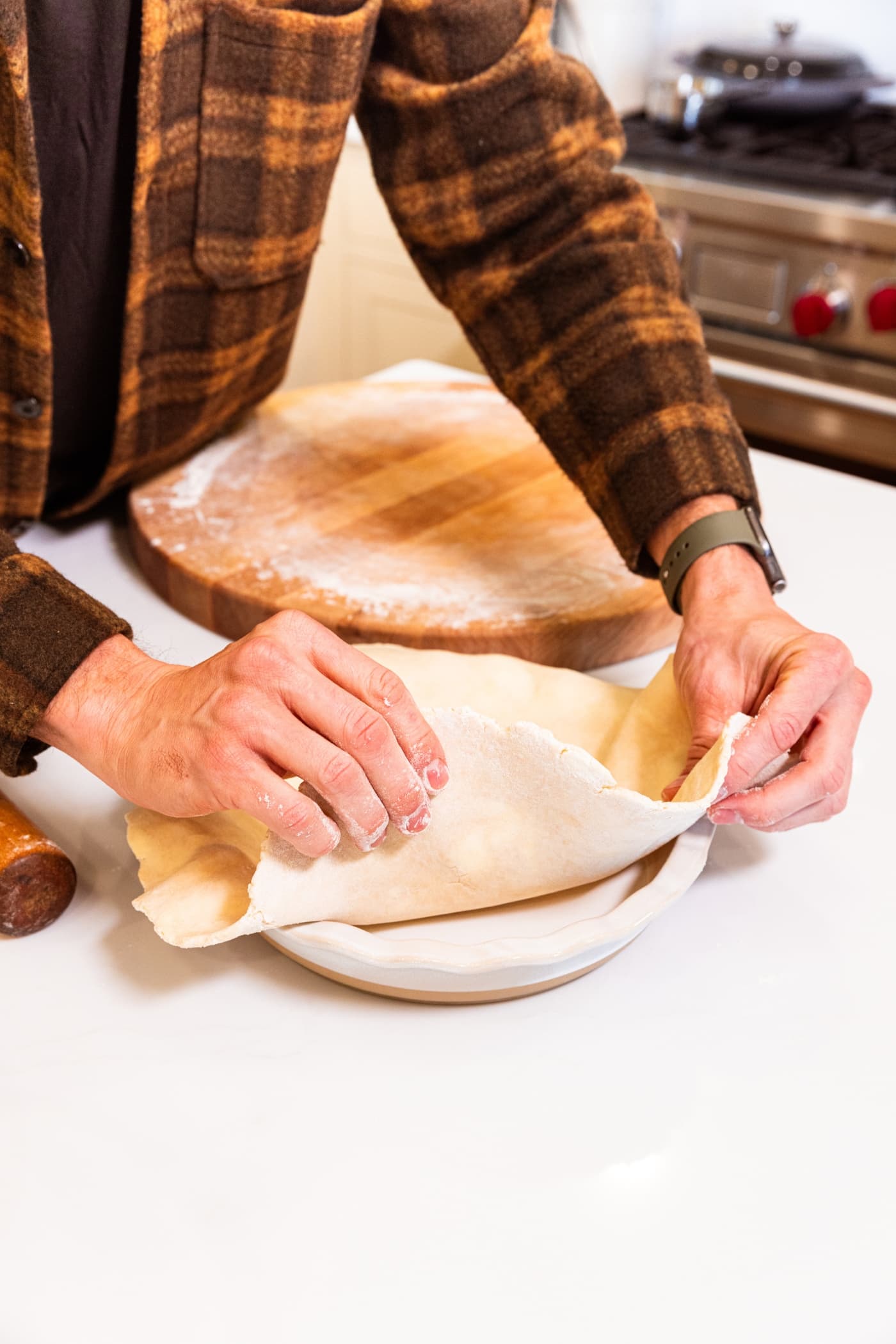 Two hands holding thin pie crust placing it into a white round pie baking dish.