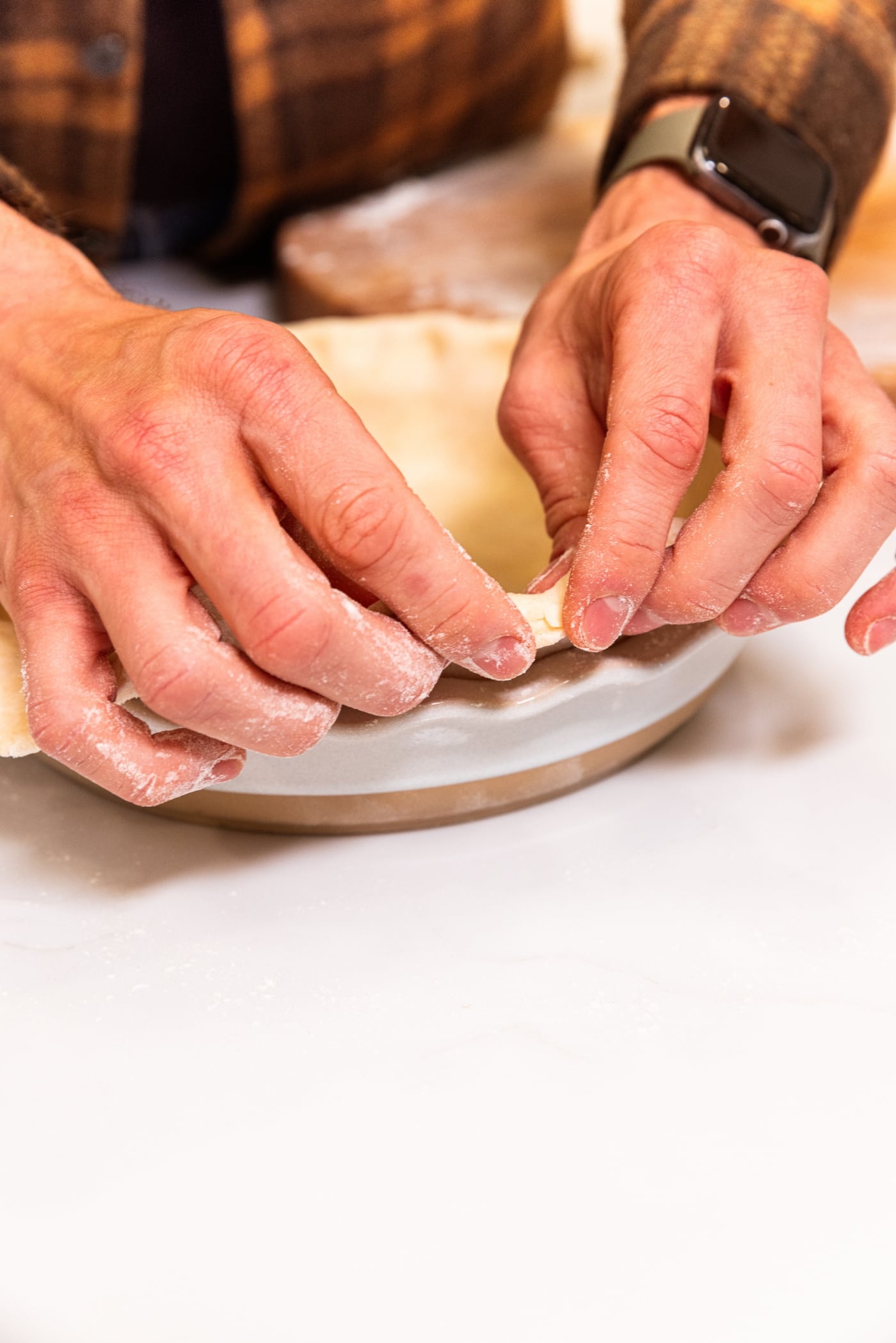 Hands folding the underside of a pie crust under in a white baking dish.