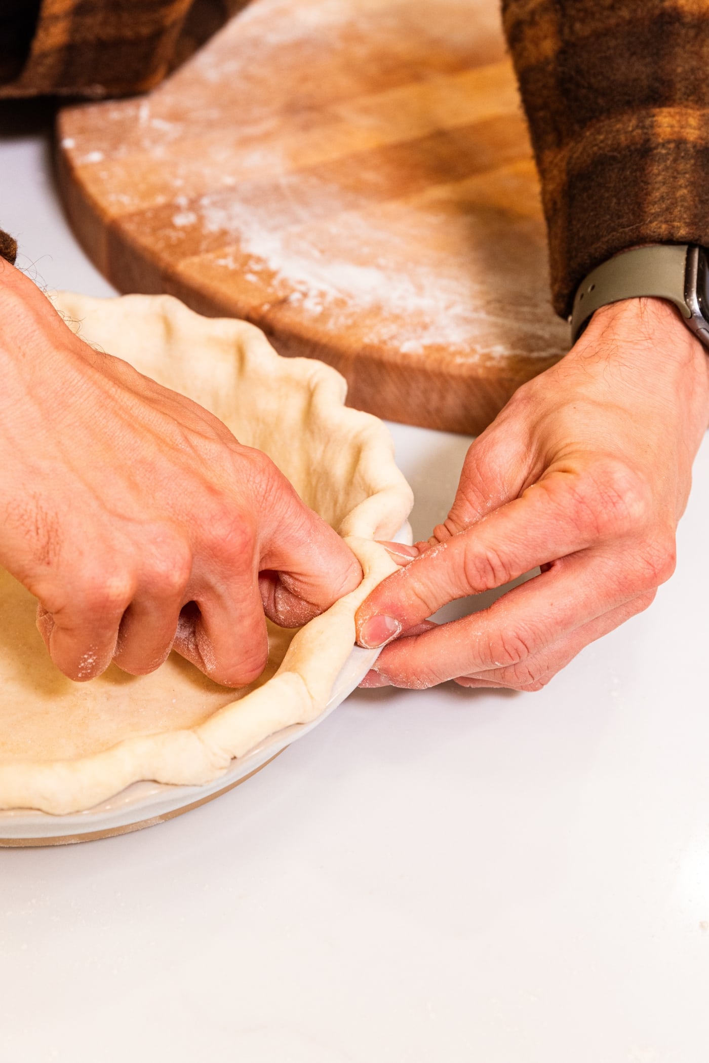 Two hands crimping the sides of a pie dough into a large white pie baking dish.