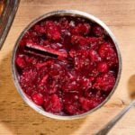 Wood surface with silver bowl filled with cranberry marmalade with cinnamon stick and silver spoon to the side.