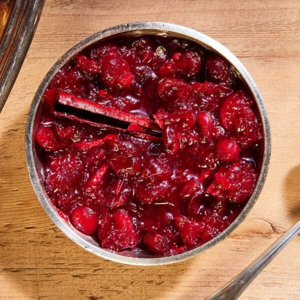 Wood surface with silver bowl filled with cranberry marmalade with cinnamon stick and silver spoon to the side.