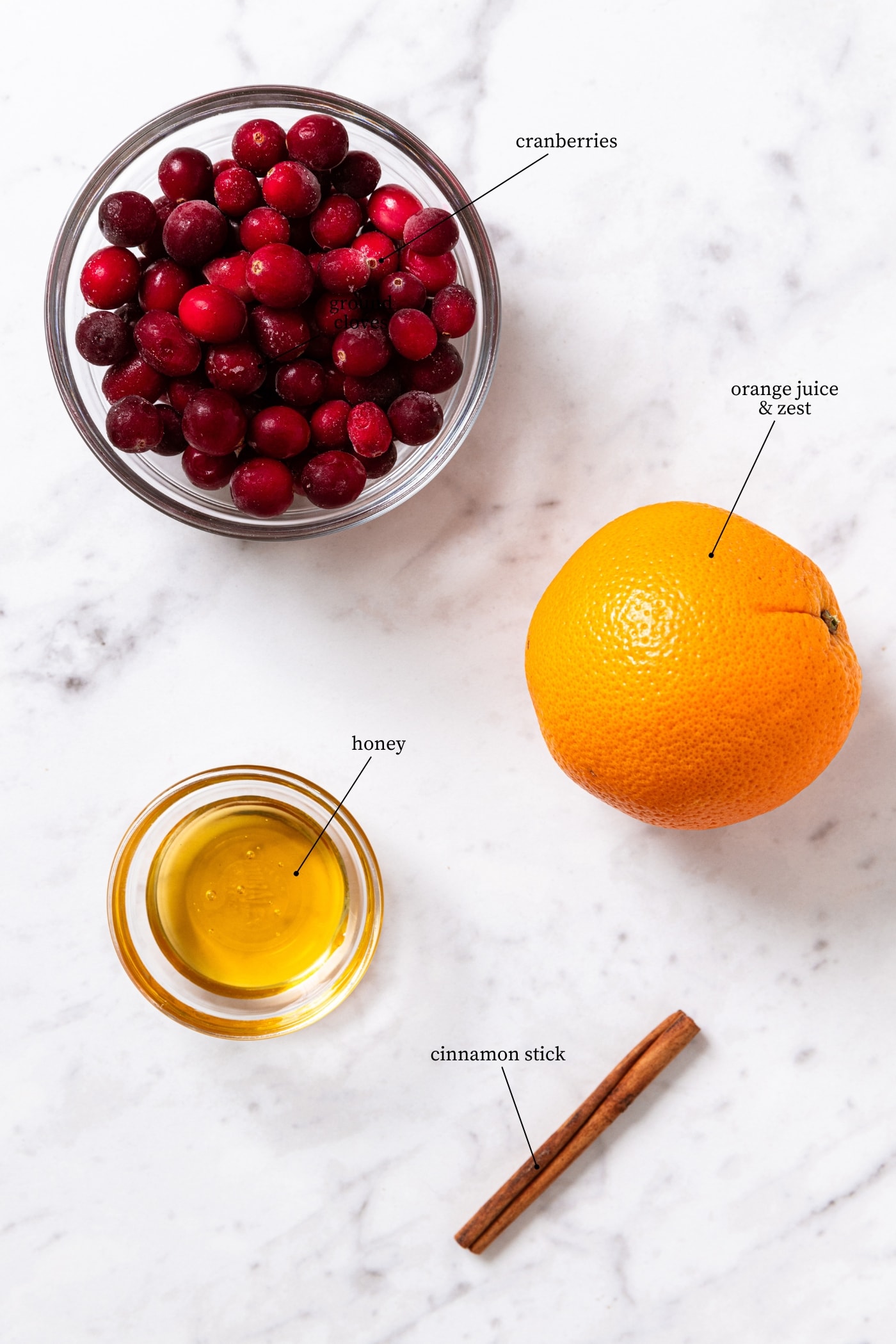 White marble surface with ingredients needed to make cranberry marmalade including cranberries, orange, honey, and cinnamon.