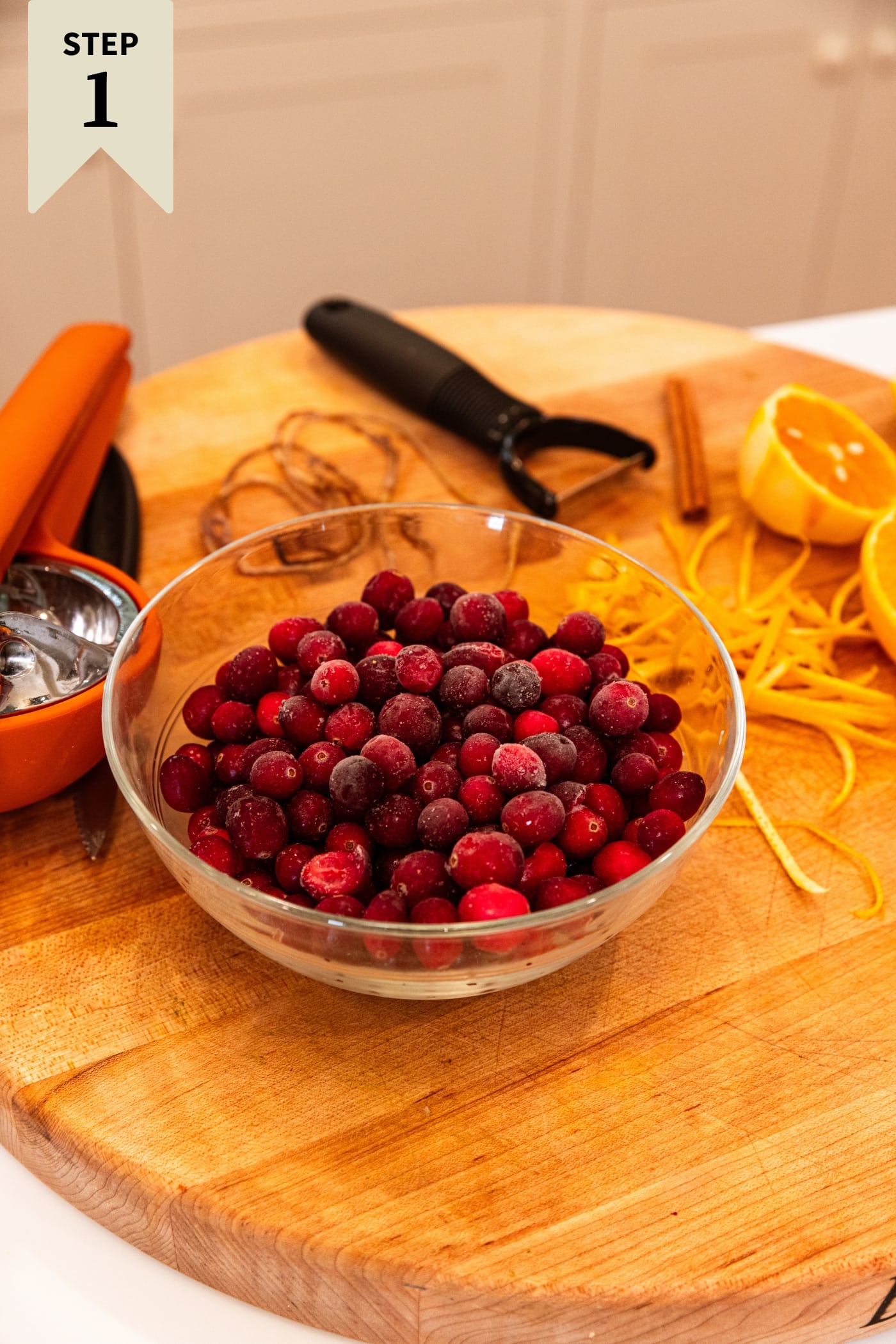 Glass bowl filled with cranberries sitting on wood cutting board with peeler in background.