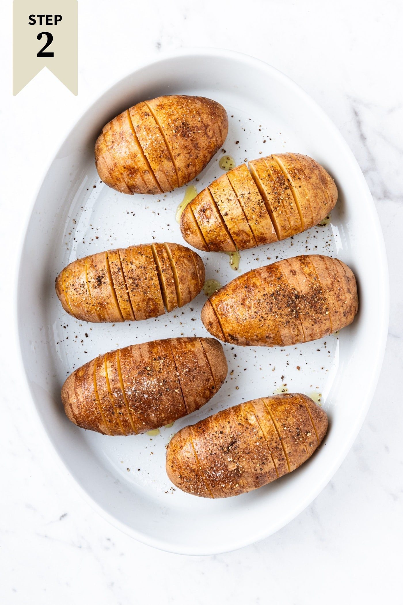 White baking dish on a marble surface with Hasselbacked potatoes sprinkled with salt and pepper before being roasted.