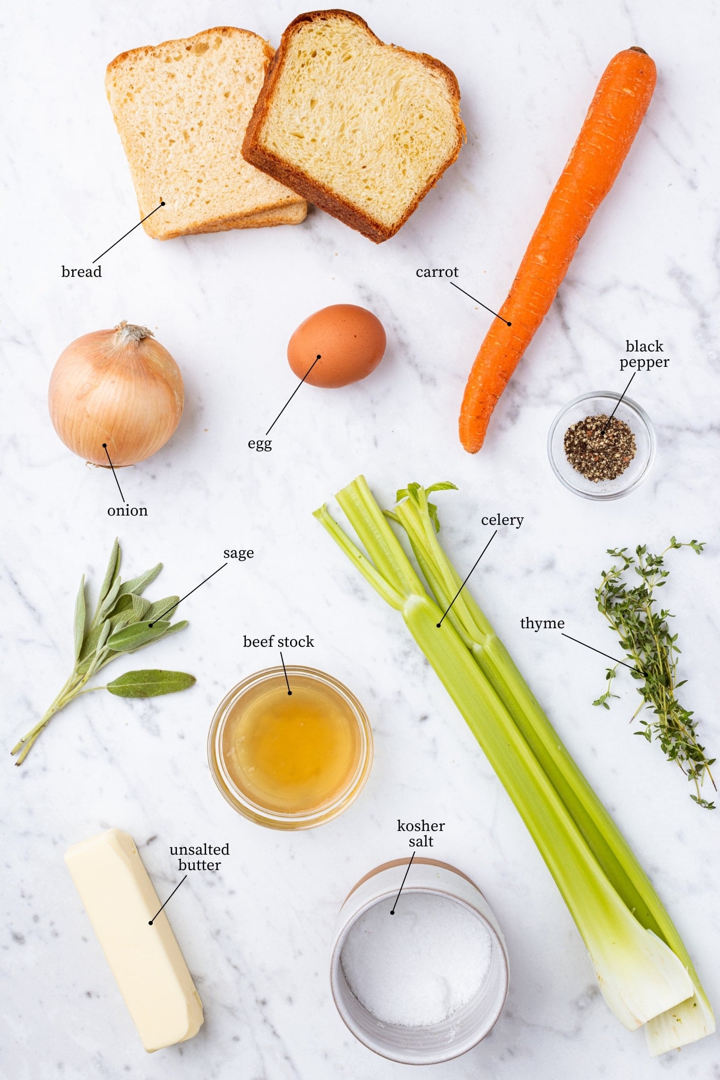 White marble surface filled with all ingredients needed to make traditional stuffing including carrots, celery, bread, onion, spices, herbs, and more.
