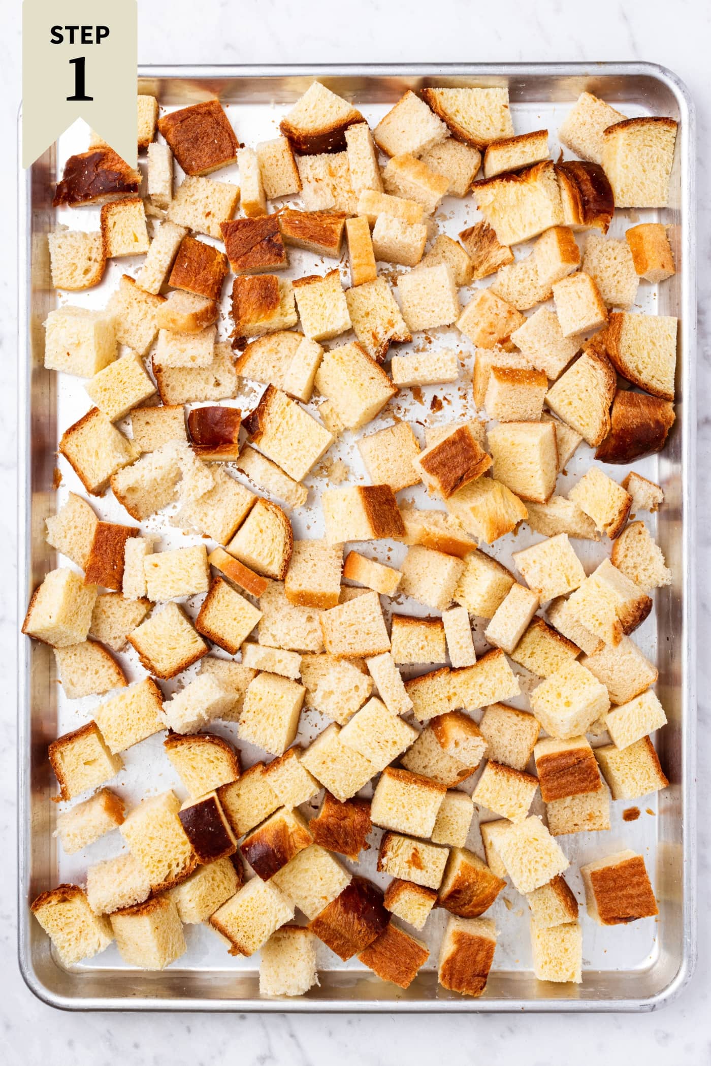 Cubes of sliced bread spread out into an even layer on a metal baking pan before toasting in the oven.