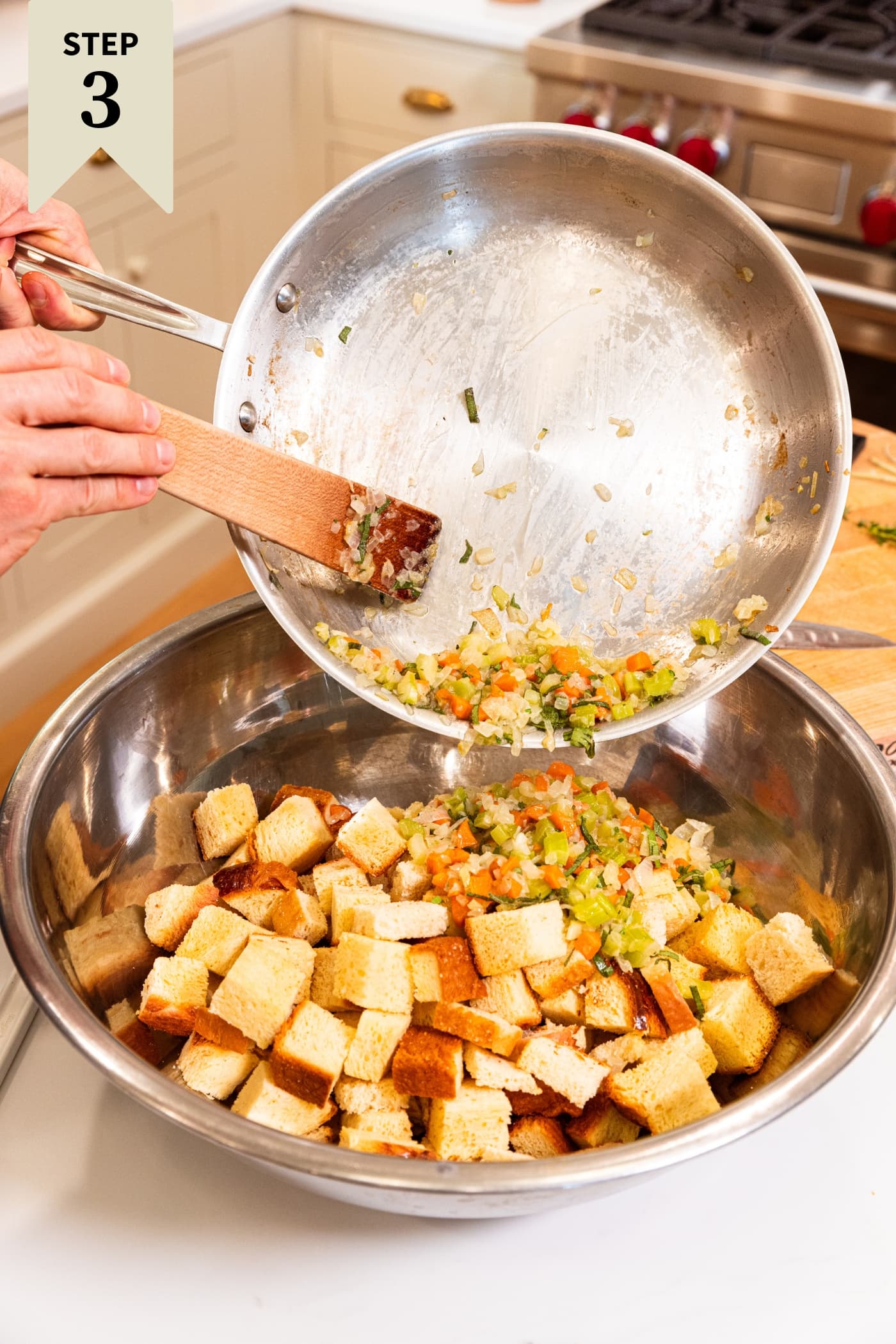 Hand holding metal skillet pouring cooked vegetables into a large metal bowl filled with toasted cubes of bread.