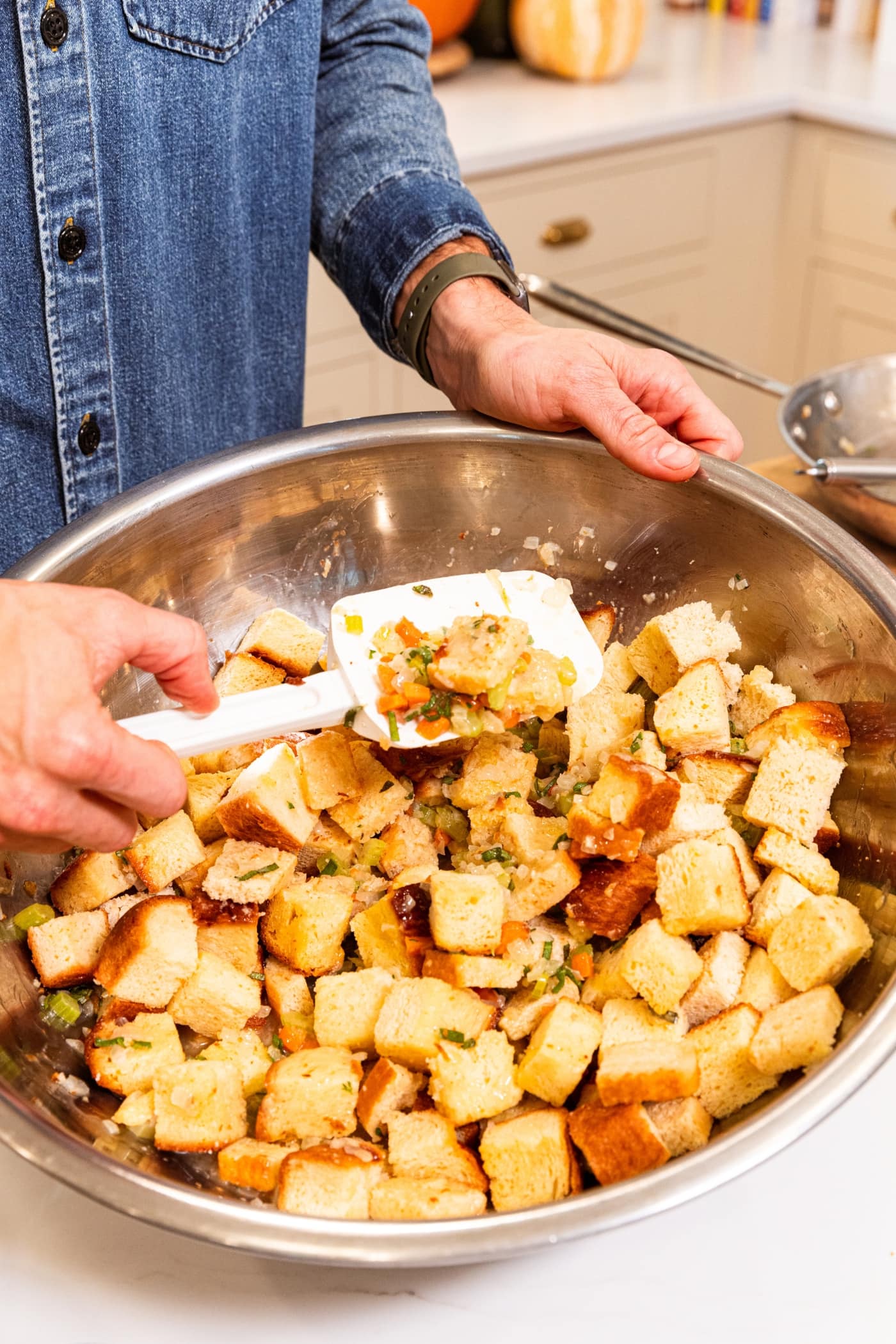 Plastic spatula mixing together bread cubes with cooked vegetables in large metal bowl for stuffing.