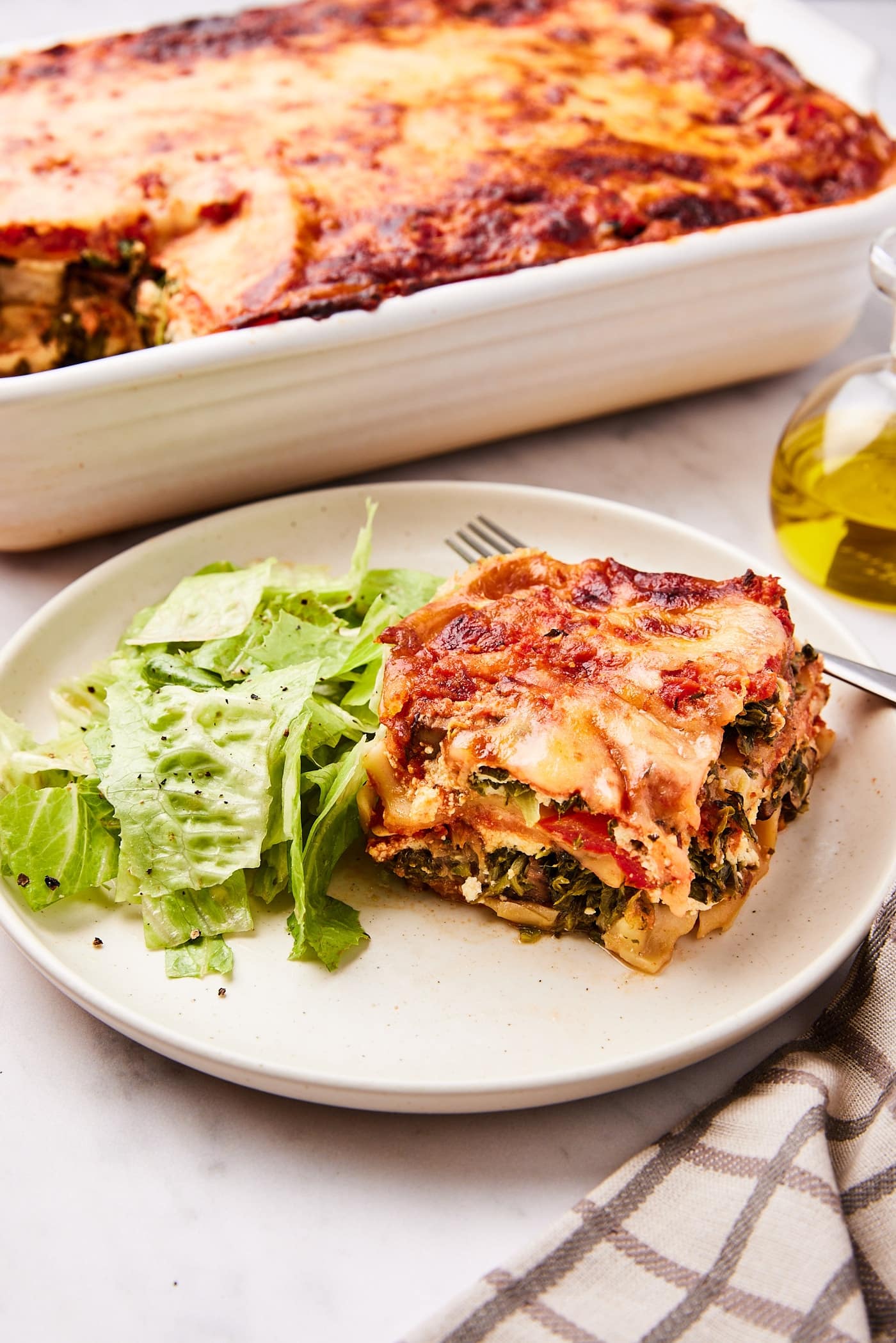 White plate with square of vegetable lasagna removed from the baking pan in the background along with a small side salad.