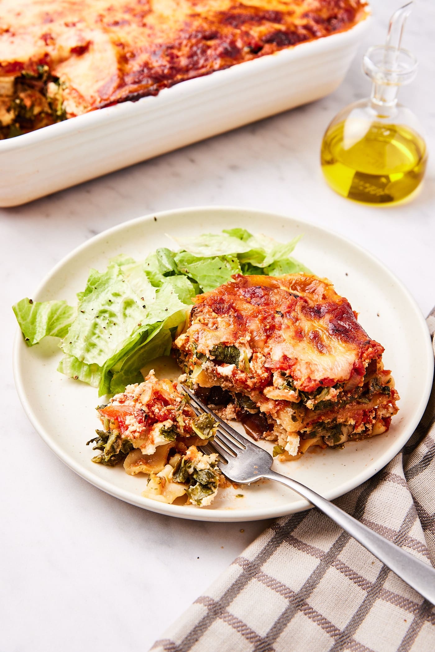 White plate with square of lasagna with side salad also on plate with rest of lasagna in baking dish behind.