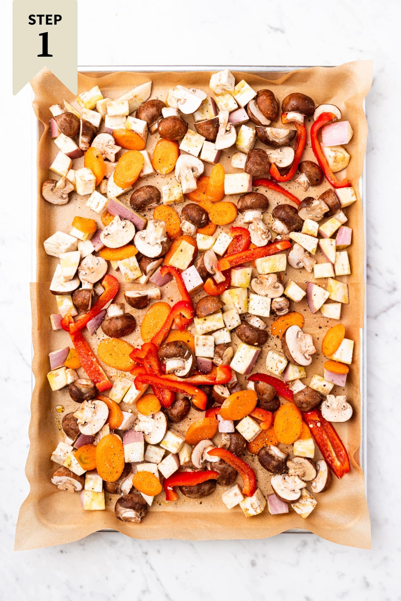 White marble surface with baking sheet with raw sliced vegetables before being put in the oven to roast.