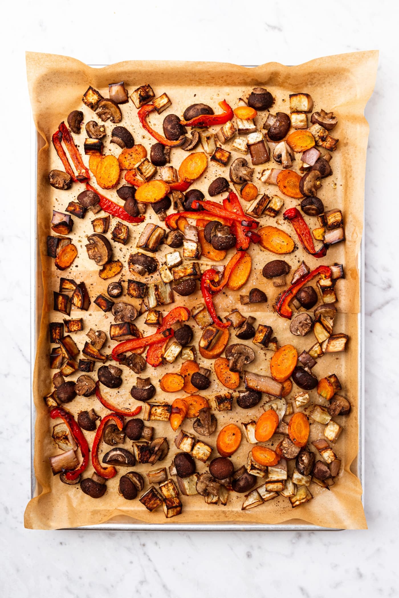 White marble surface filled with parchment-lined baking sheet filled with roasted vegetables after coming out of the oven.