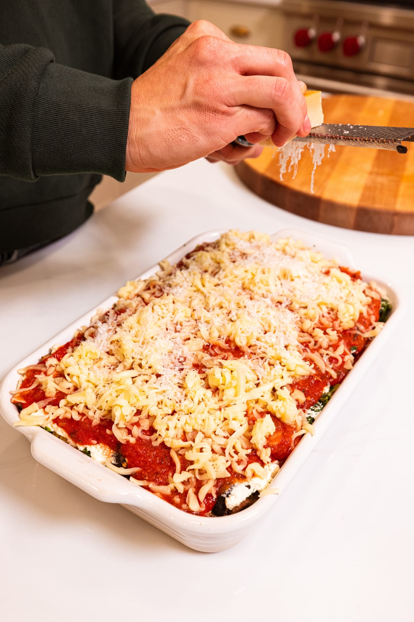 Hand holding grater spreading fresh Parmesan cheese over the top of an assembled lasagna in a baking dish.