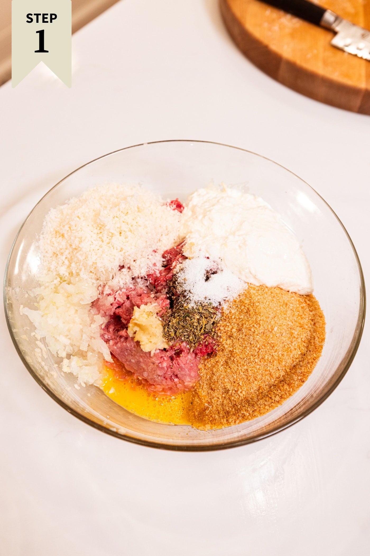 Glass bowl filled with meatball ingredients before being mixed together all on white surface.