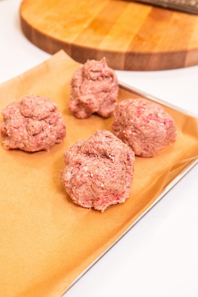 Round balls of meat mixture sitting on parchment-lined baking sheet on white surface to form meatballs.
