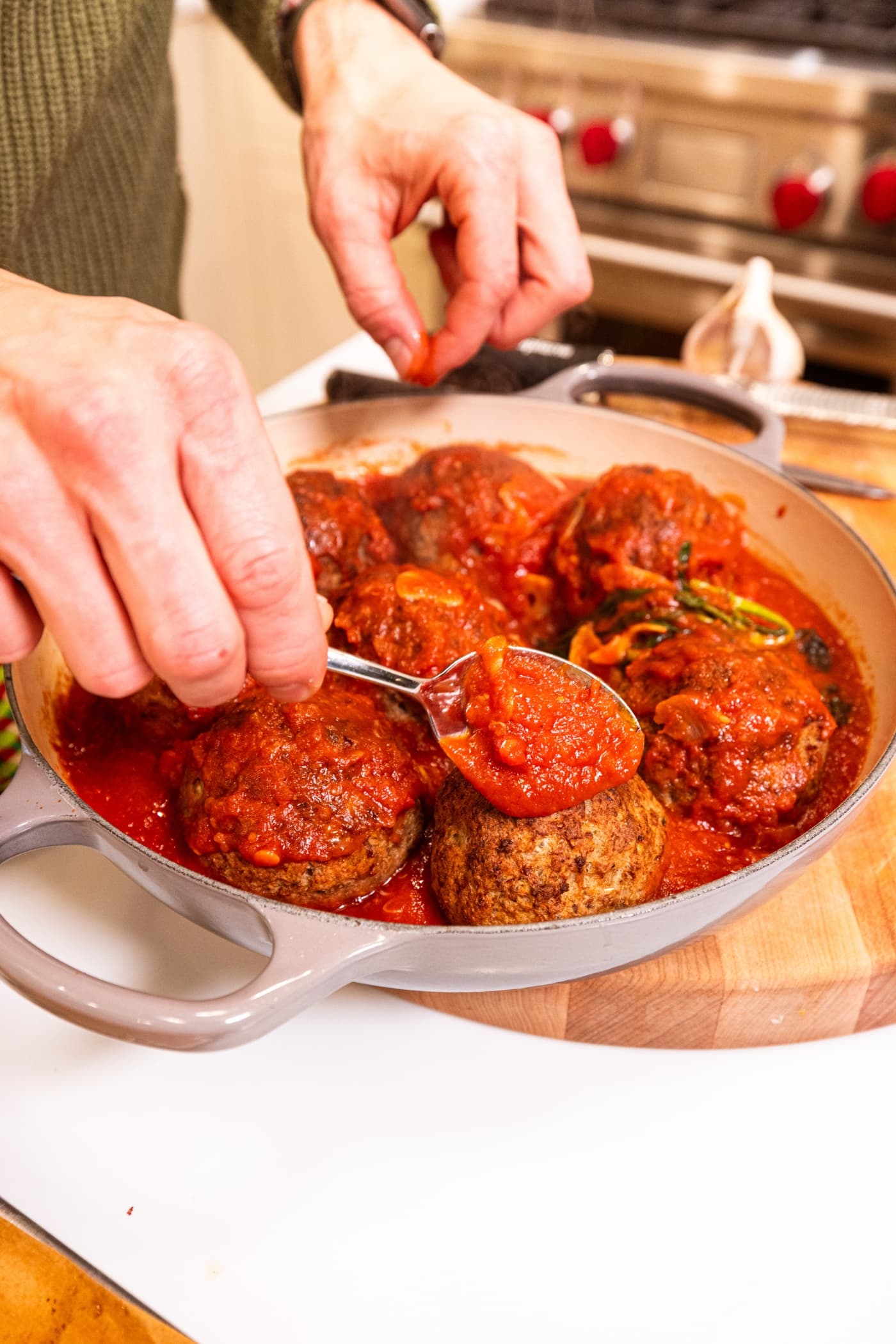 Hand using spoon to pour red sauce mixture over the top of meatballs in a large round baking dish.