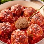 Round baking dish filled with meatballs topped with red tomato sauce with center one sliced in half showing interior texture.