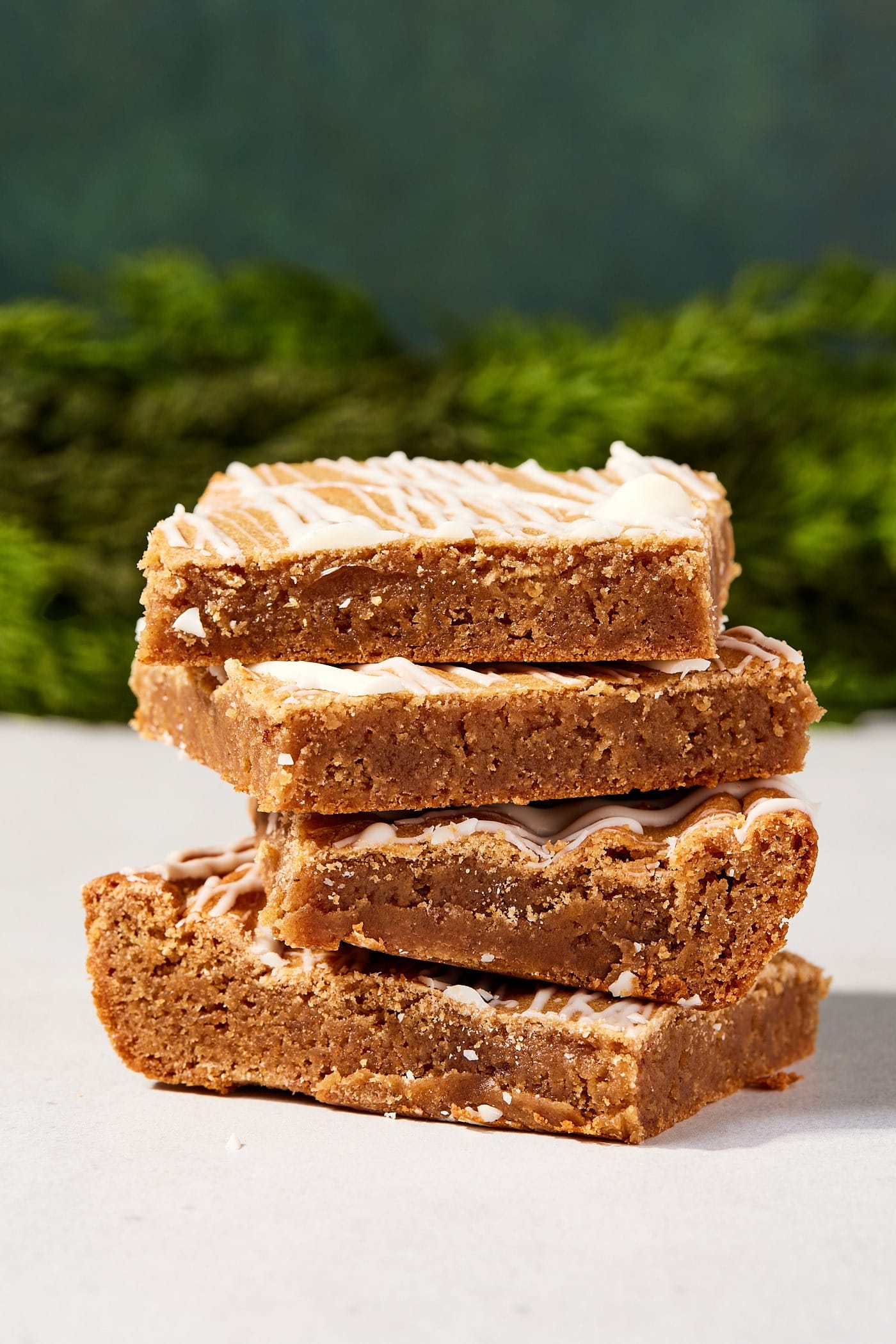 Stack of four gingerbread blondies with melted chocolate on top sitting on white surface with holiday greenery in the background.