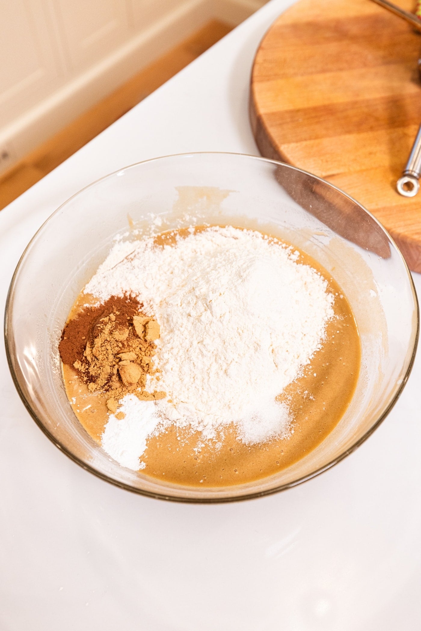 Glass bowl filled with wet ingredients with dry ingredients and spices on top all on white surface.
