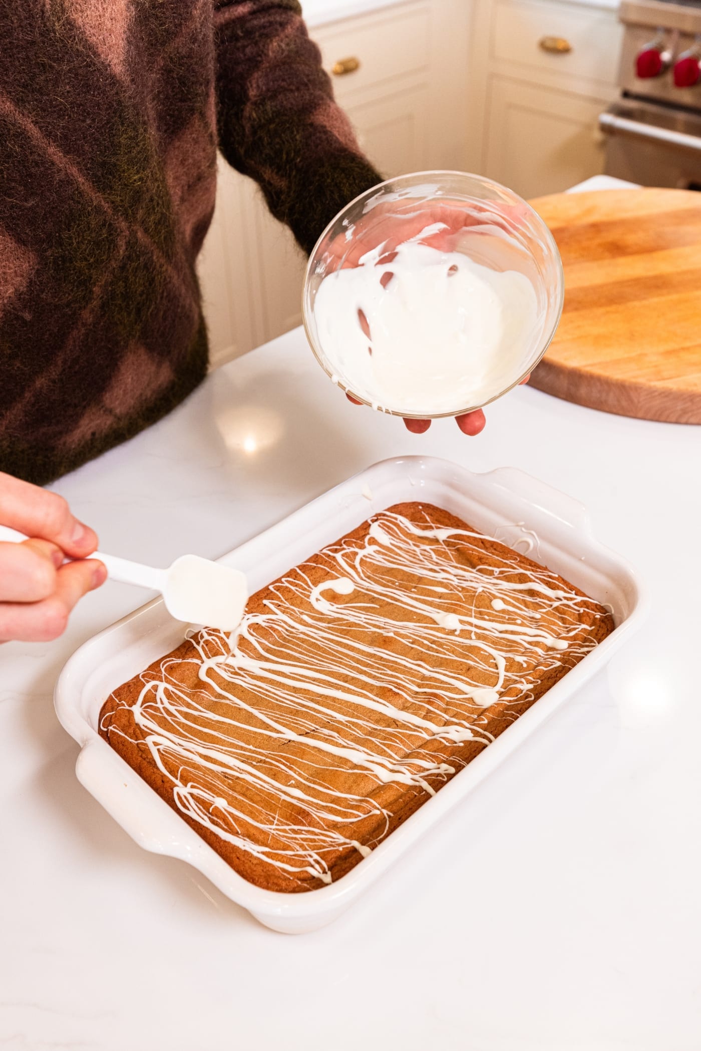 Hand using spatula to drizzle melted white chocolate over the top of gingerbread blondies in a white baking dish.