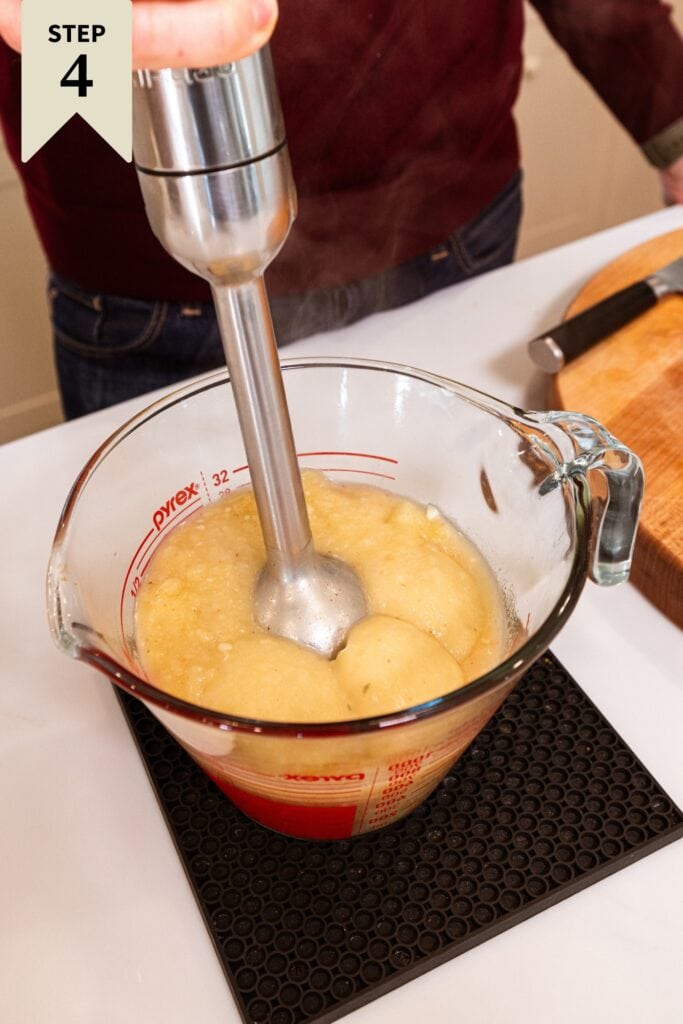 Hand using immersion blender to purée pieces of potato in potato soup to be creamy.