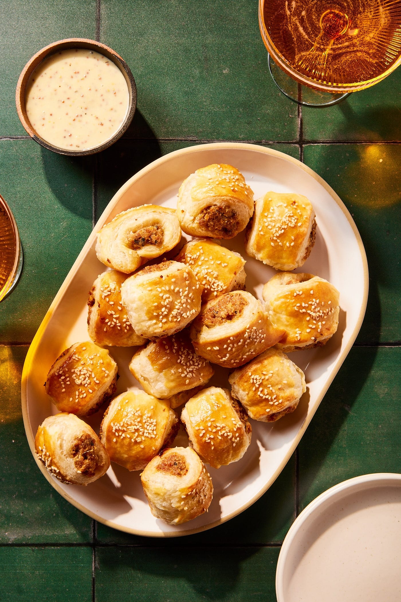 Green tiles with white platter with puff pastry sausage bites freshly baked in the oven with bowl of mustard sauce.