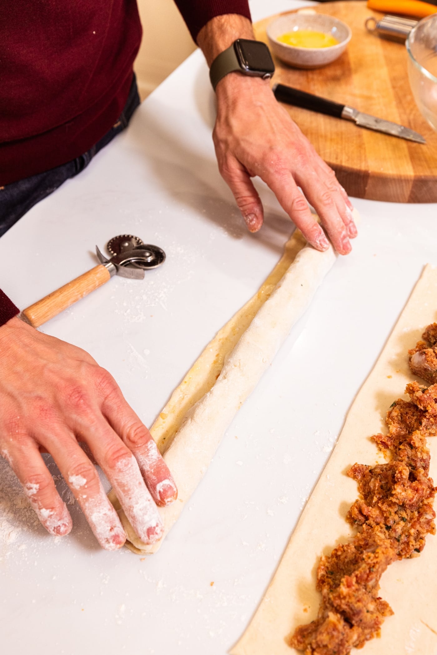 Two hands rolling puff pastry into a log filled with sausage mixture.