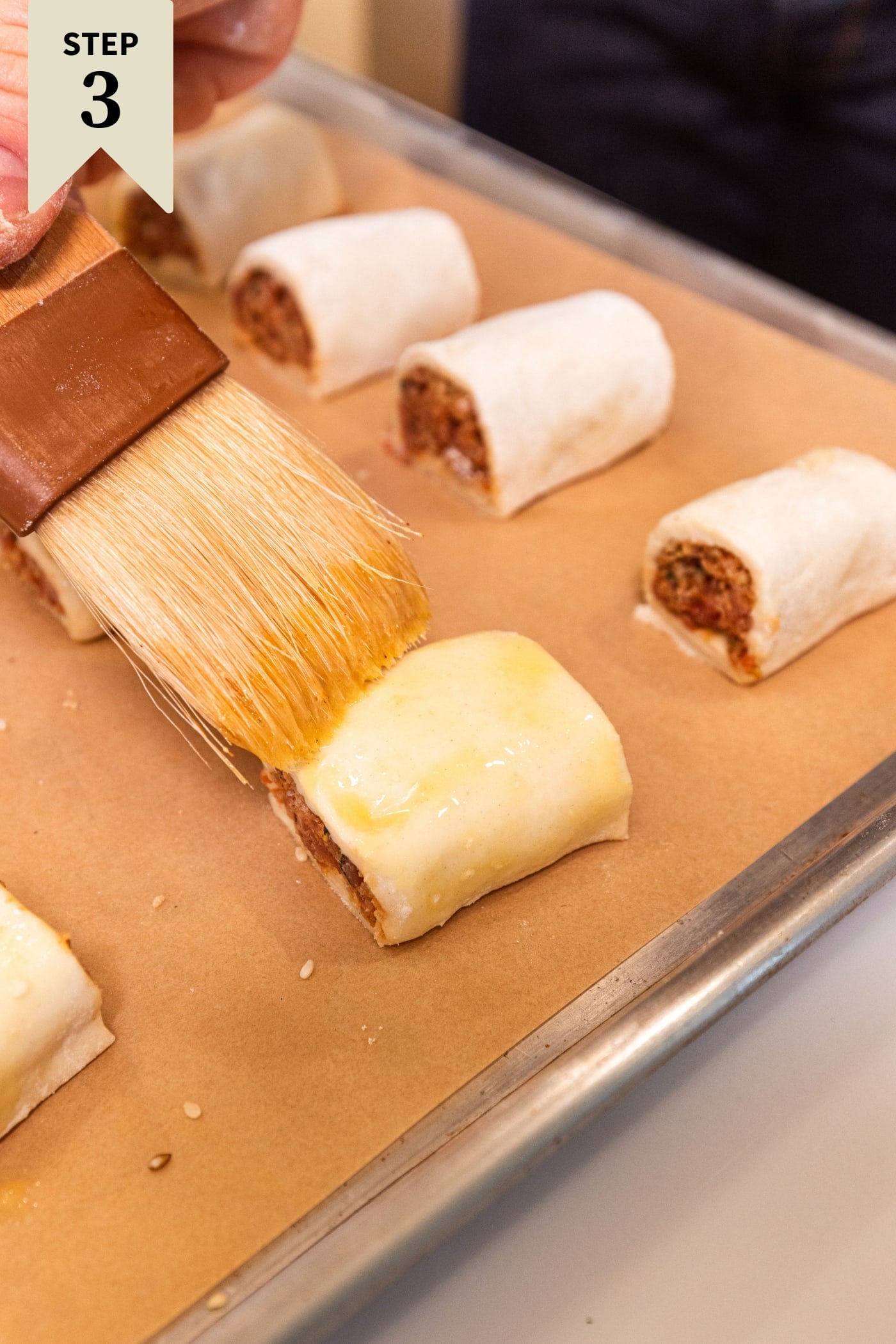 Brush brushing on egg wash on top of puff pastry sausage bites on parchment-lined baking sheet.