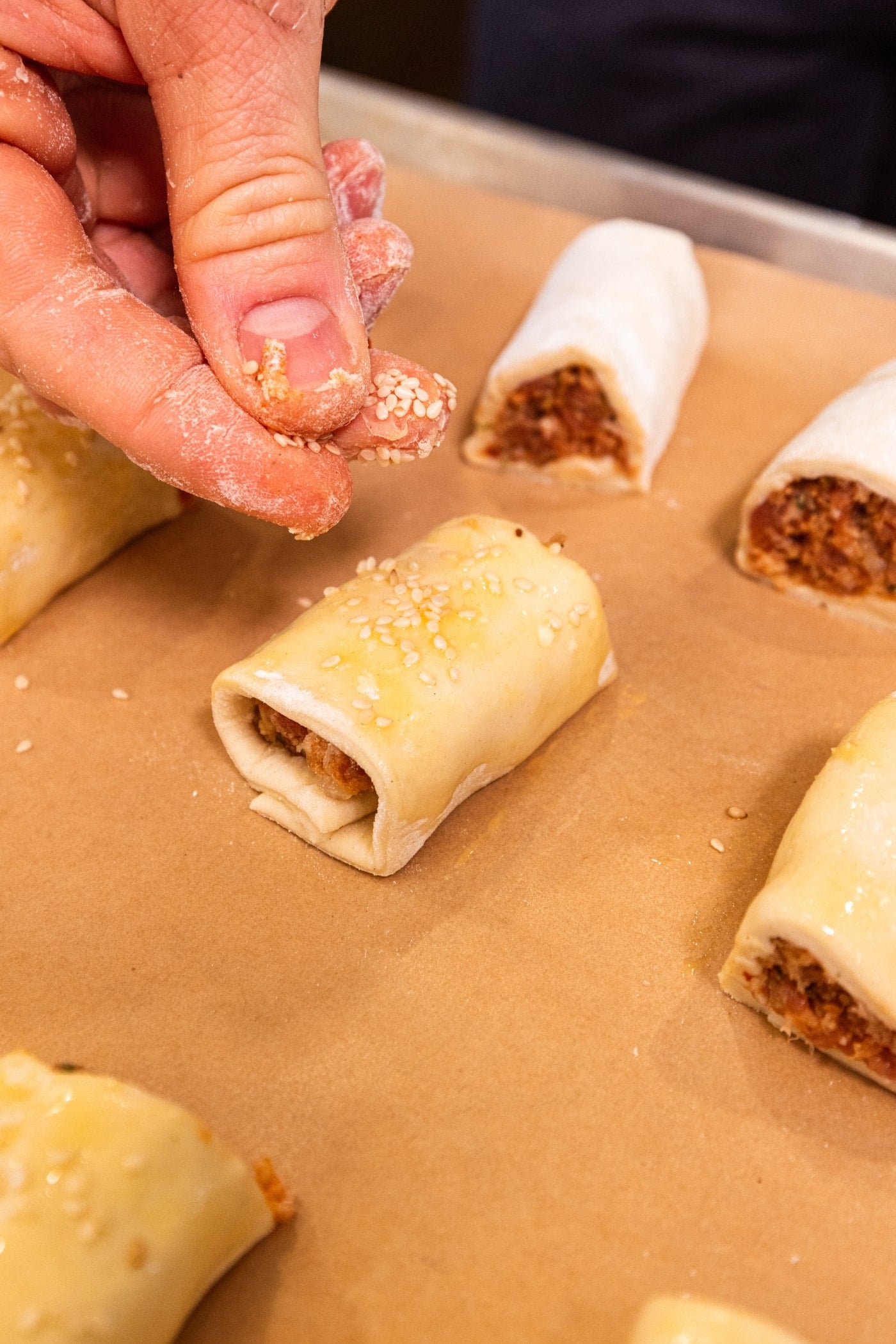 Hand sprinkling sesame seeds on top of puff pastry bites on piece of parchment.