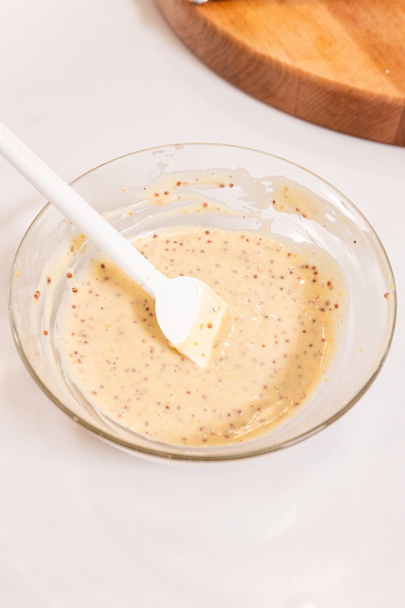 Mustard sauce with stone ground pieces in glass bowl after being mixed together with spatula leaning against side of bowl.