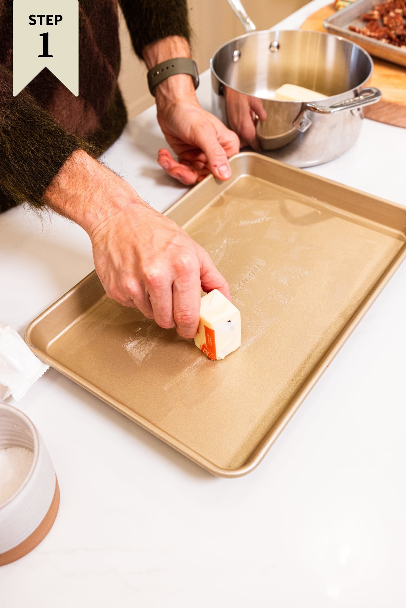 Hand using a stick of butter rubbing it into the bottom of a gold pan to create a greased layer.