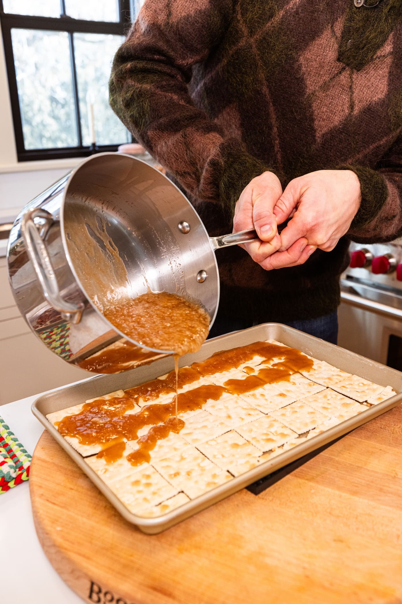 Two hands pouring caramel topping on top of saltine crackers in a gold baking pan from a saucepan hot from the stove.