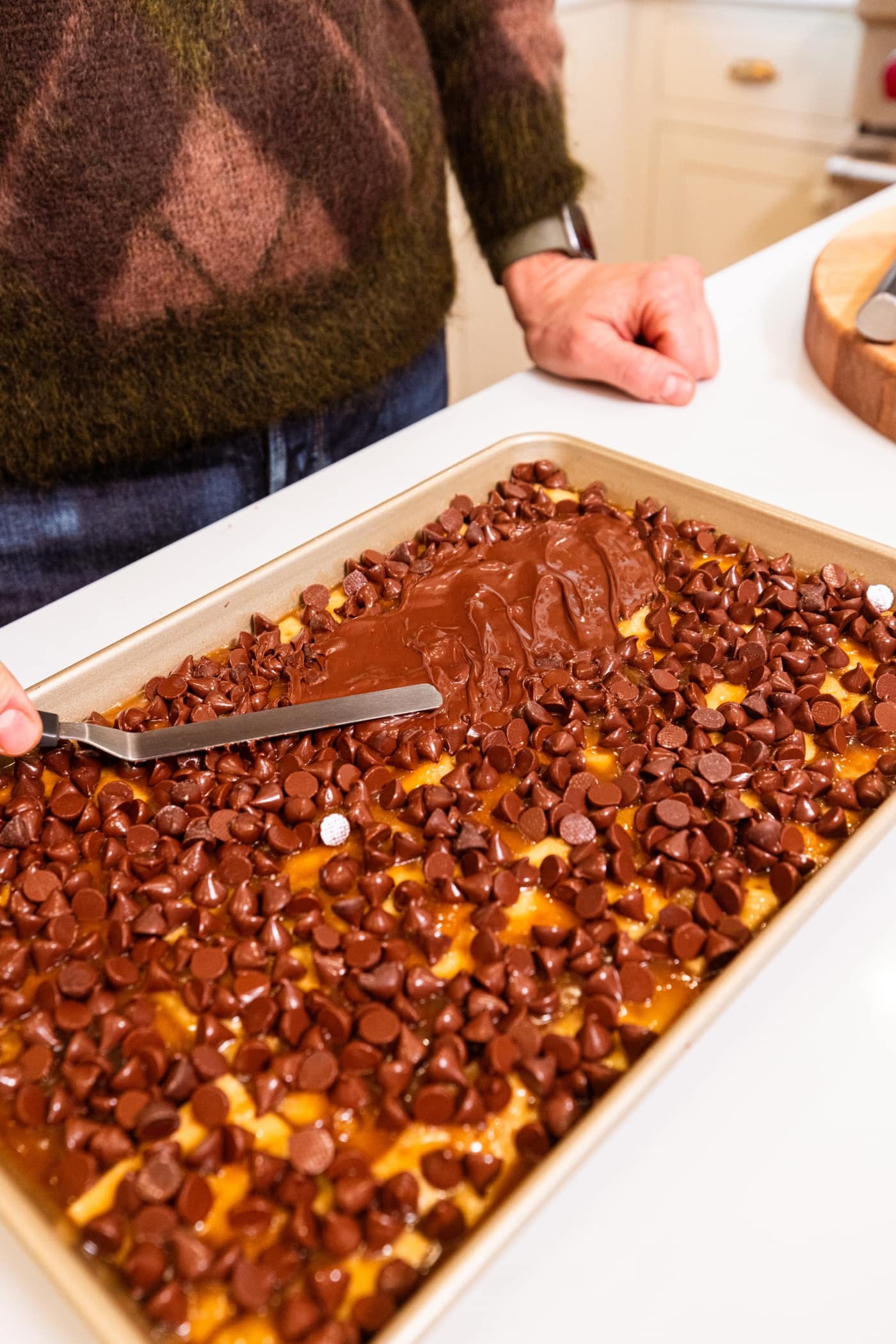 Hand using offset spatula to smooth melting chocolate chips over the top of a caramel layer for saltine toffee.