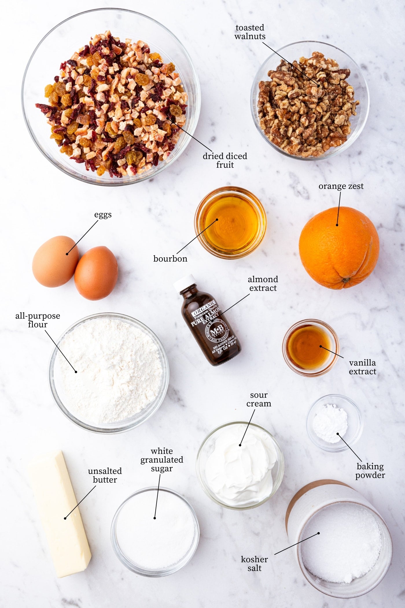White marble surface filled with all ingredients needed to make white fruitcake including yuletide mix, orange, eggs, dry ingredients, and more.