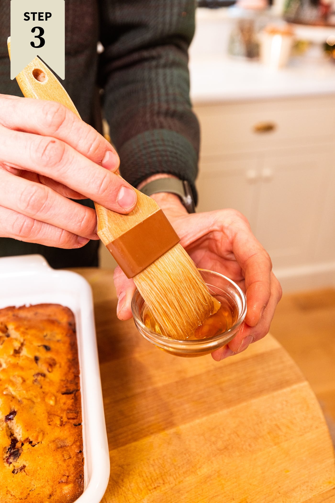 Hand using brush to dip into a small bowl filled with bourbon before brushing on top of white fruitcake.
