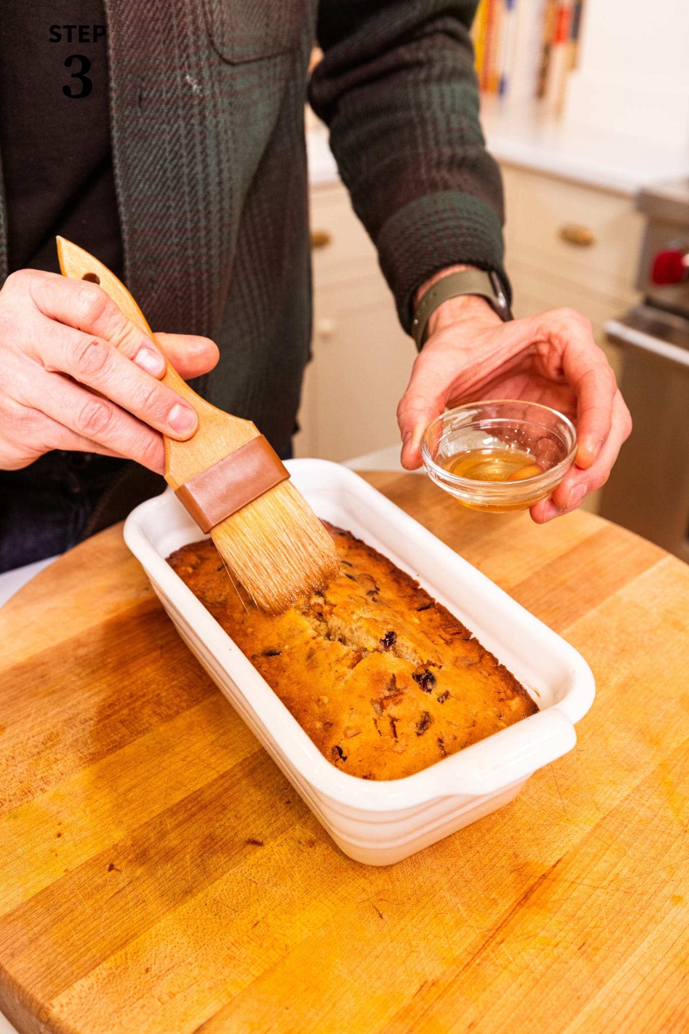 Hand using brush to brush bourbon on the top of white fruitcake after being baked in a white loaf pan.
