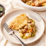 White plate resting on tablecloth with serving of chicken pot pie with fork resting on plate as well with more pot pie in dish behind.
