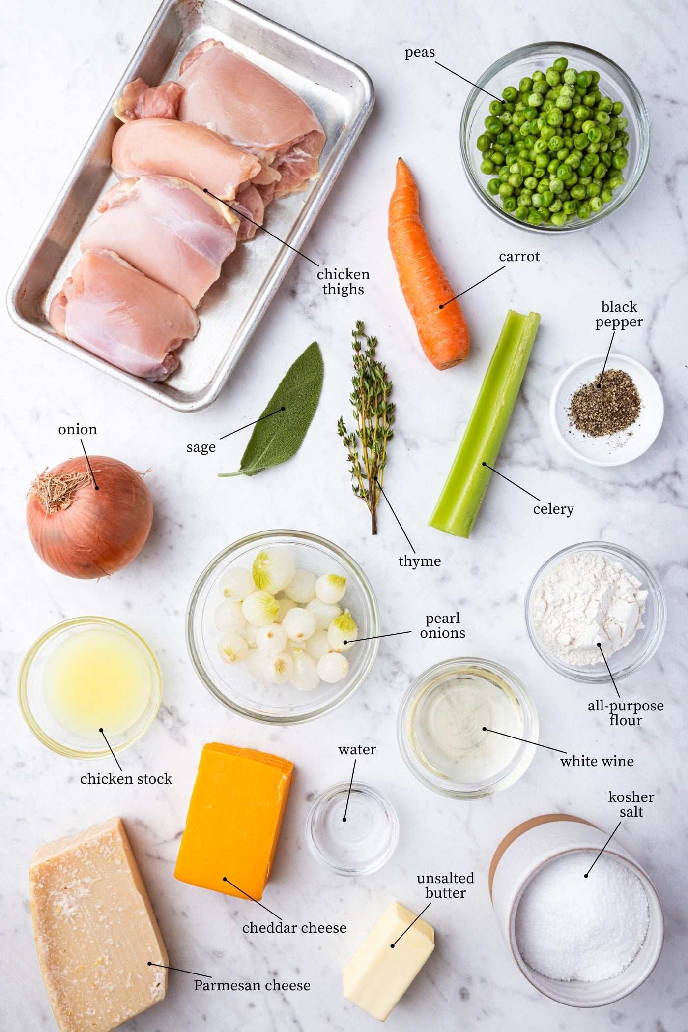 White marble surface filled with all ingredients needed to make chicken pot pie including onion, celery, carrot, raw chicken, peas, cheese, and more.