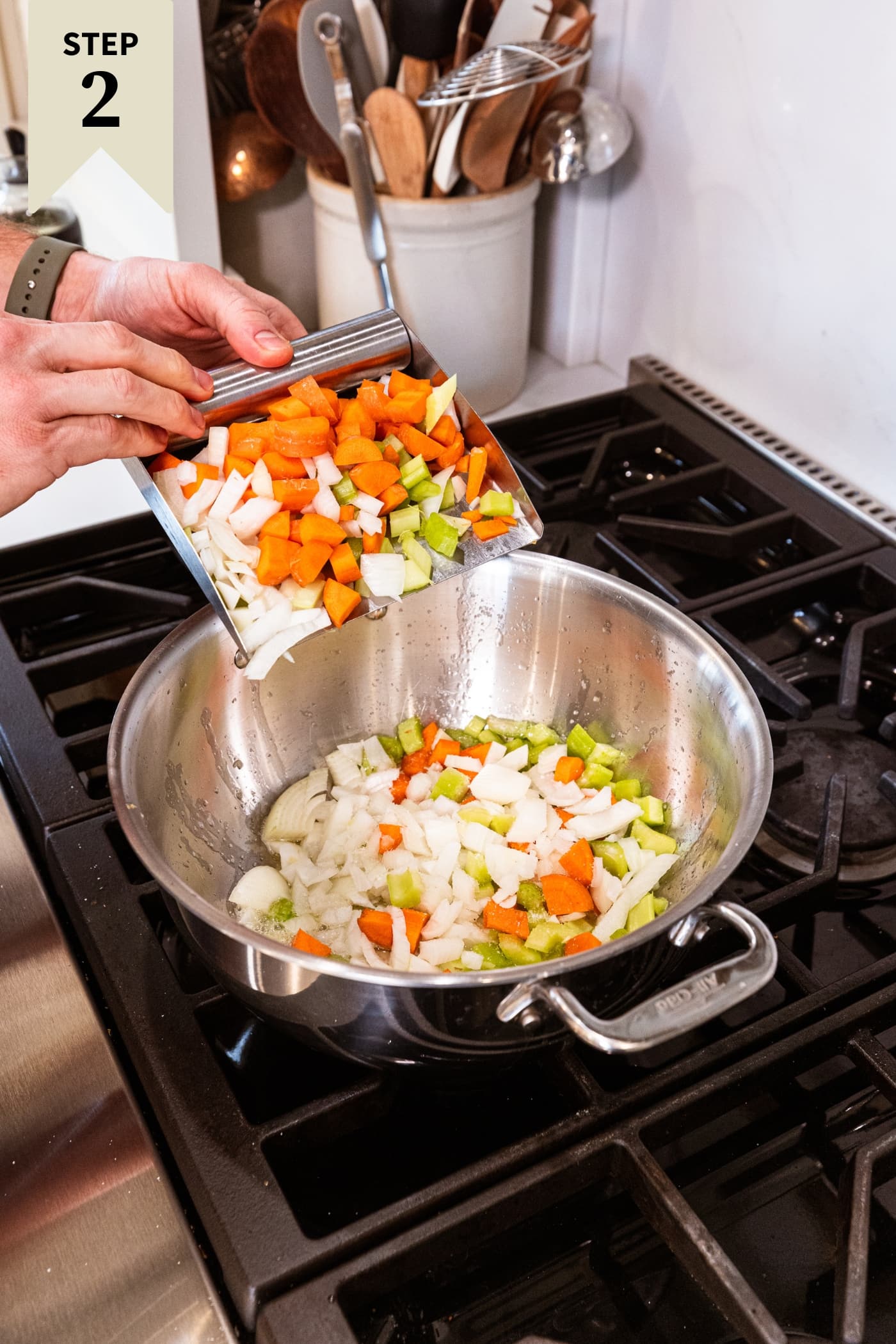 Hand scooping chopped carrot, celery, and onion into a large stockpot over the heat.