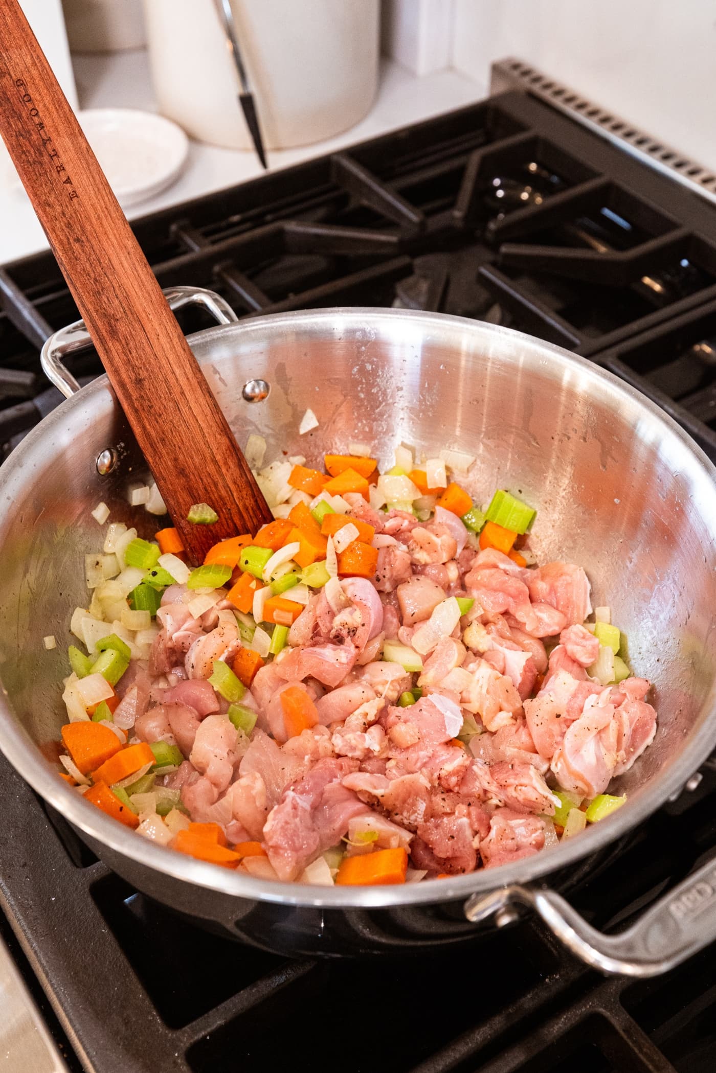 Raw chicken cooking amongst mixture of carrot, onion, and celery with wooden spatula over the heat.