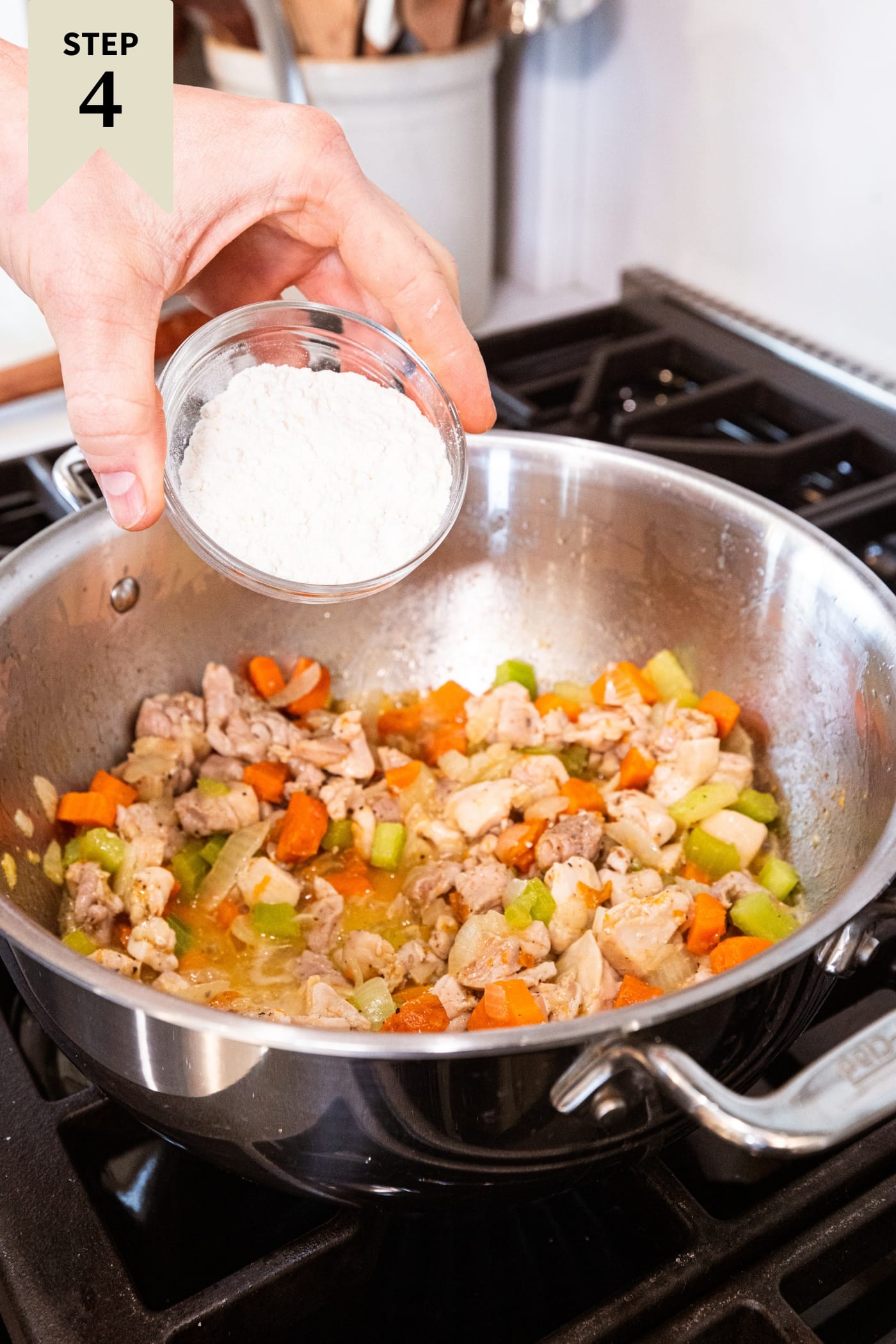 Hand holding small bowl of flour about to sprinkle on top of cooked chicken mixture in pot over the heat.