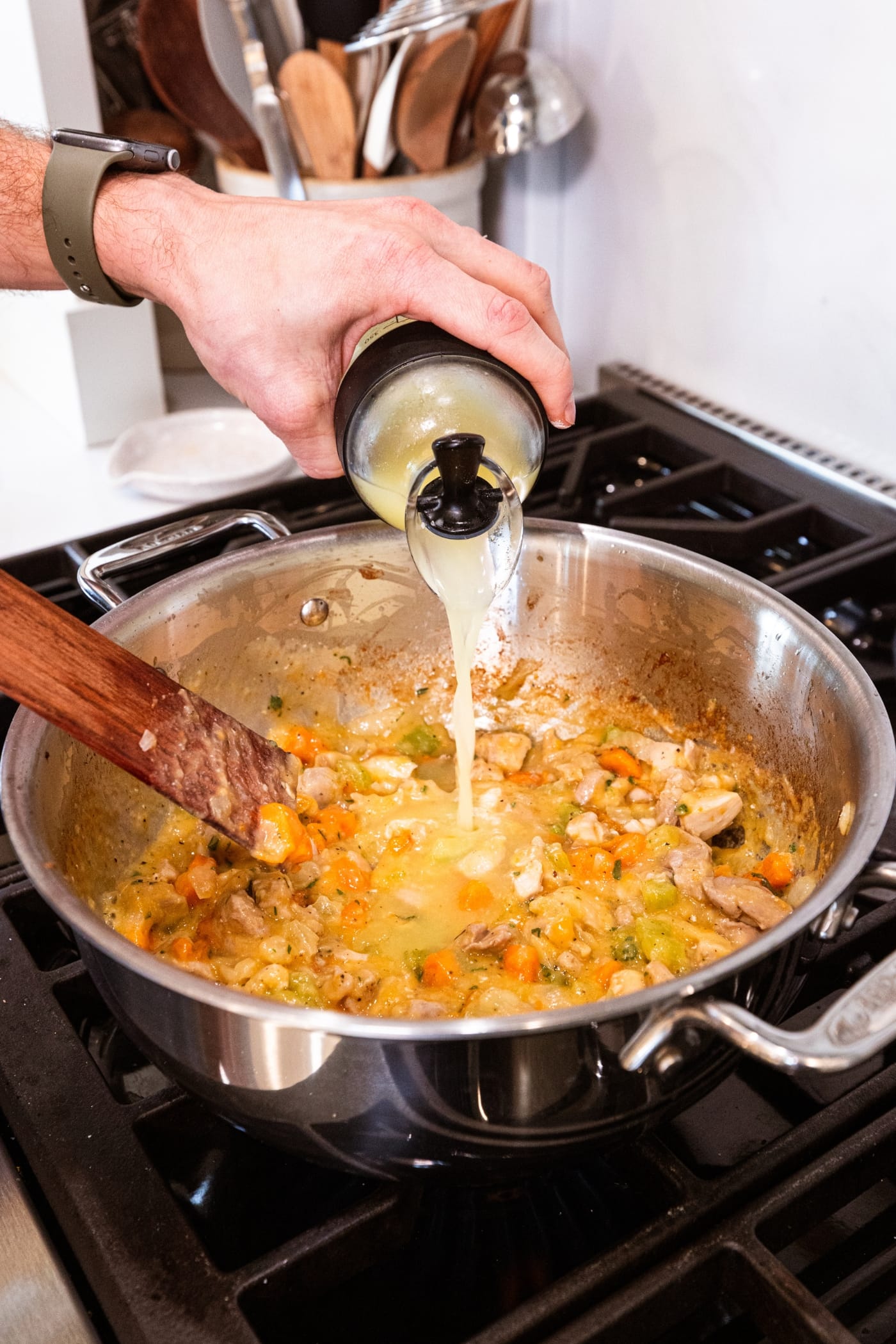 Hand pouring chicken stock into pan filled with thickened chicken mixture over the heat.