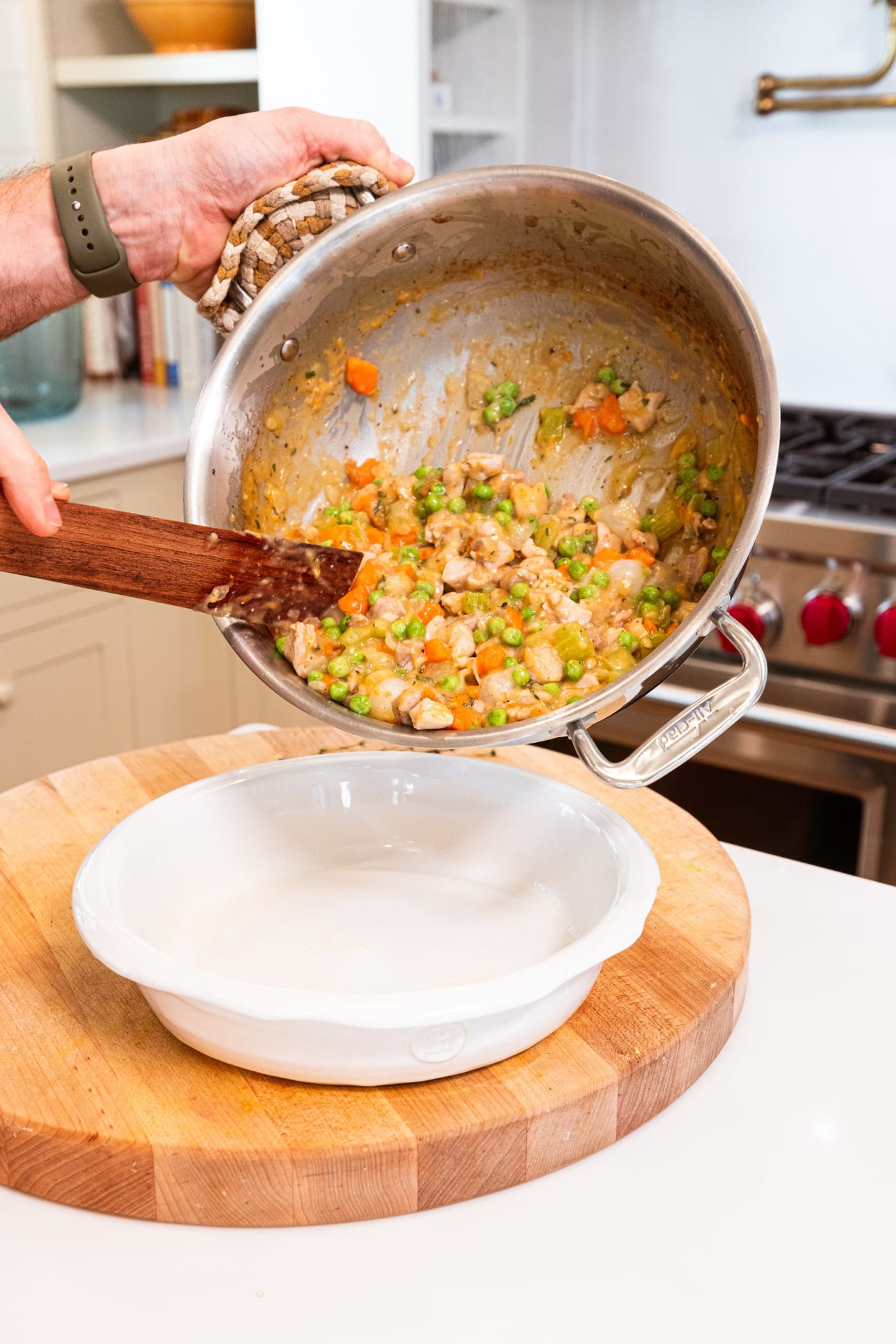 Hands about to pour chicken pot pie mixture into a round pie dish on wooden cutting board.